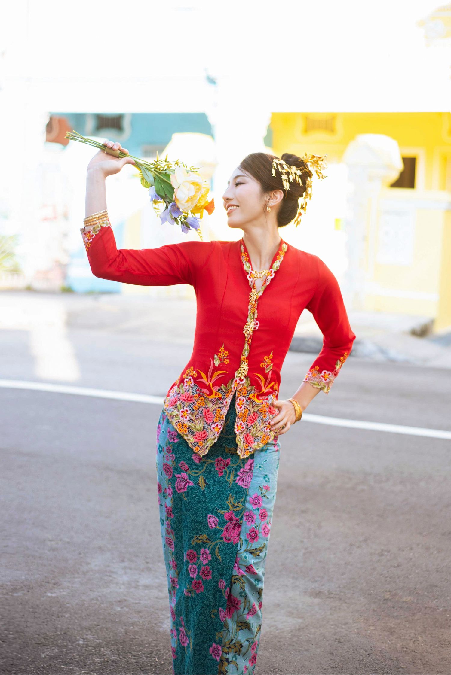 Traditional Indonesian dancer wearing red kebaya with gold embroidery and floral skirt performs graceful pose on street.