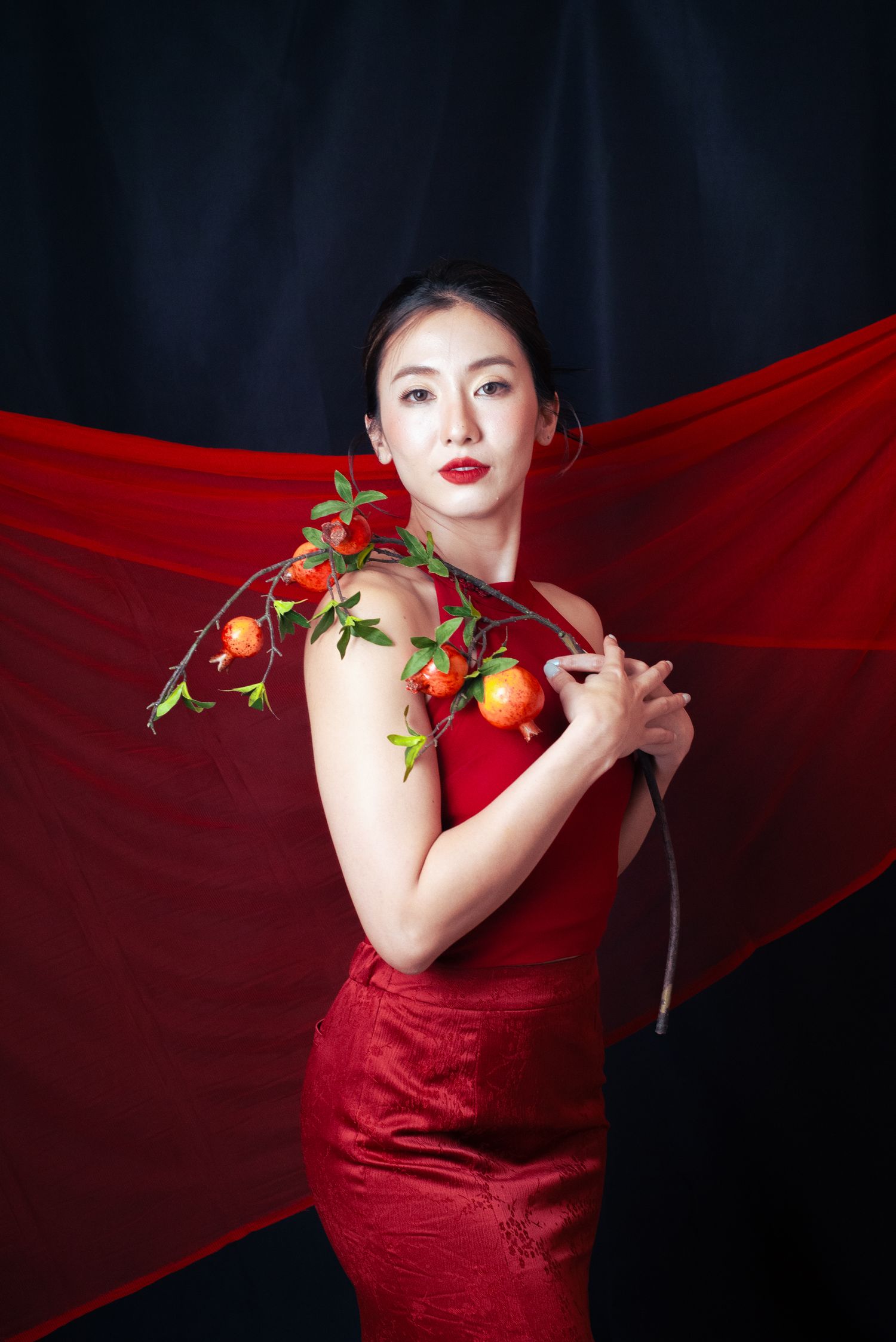 Woman in red qipao dress holding pomegranate branch with red fabric backdrop in elegant portrait.