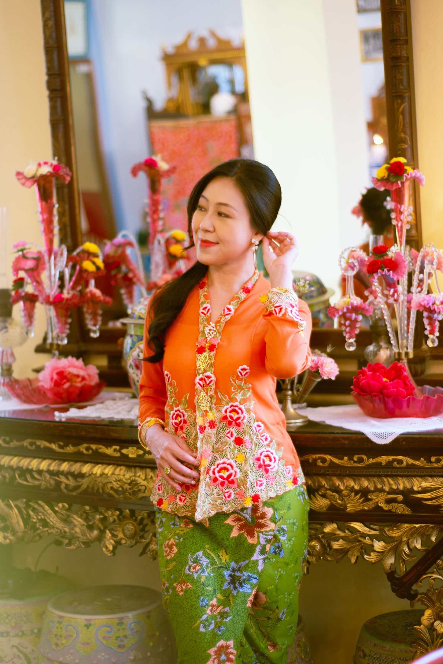 Traditional Indonesian kebaya outfit in orange and green with floral embroidery displayed in ornate indoor setting with mirrors.