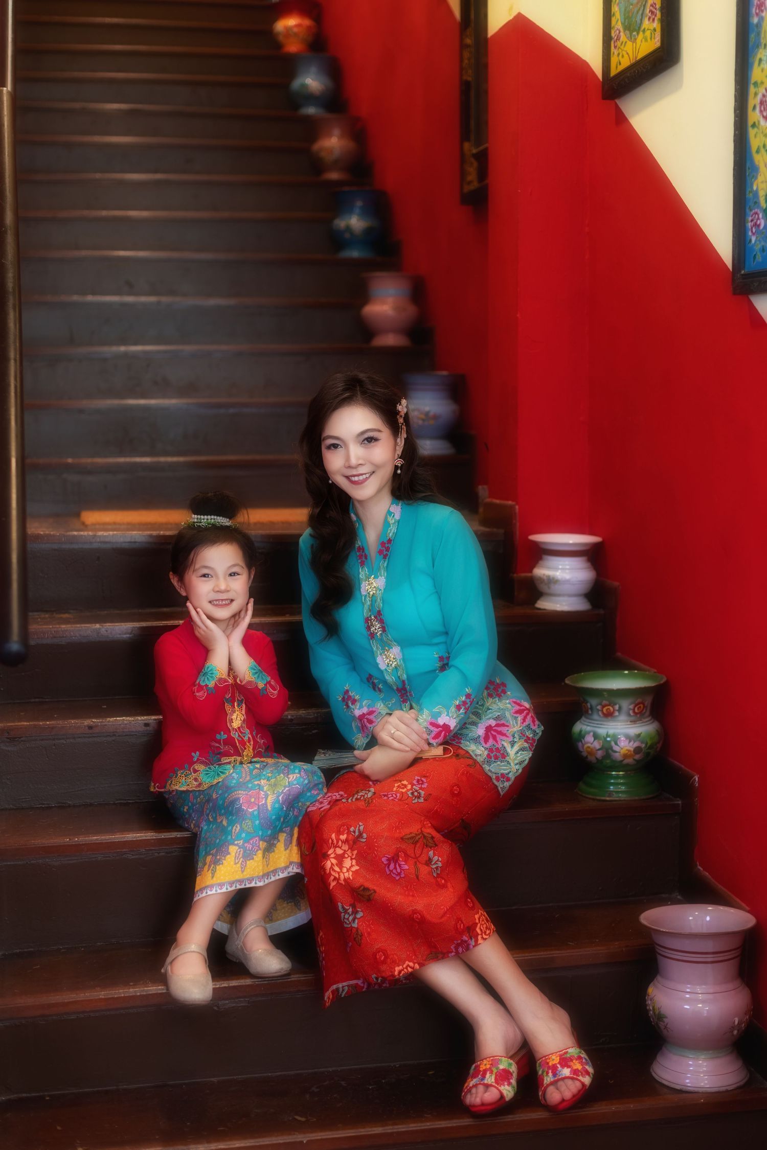 Photoshoot of a Mother and daughter in traditional Nyonya Kebaya sitting on red-walled stairs with decorative vases.