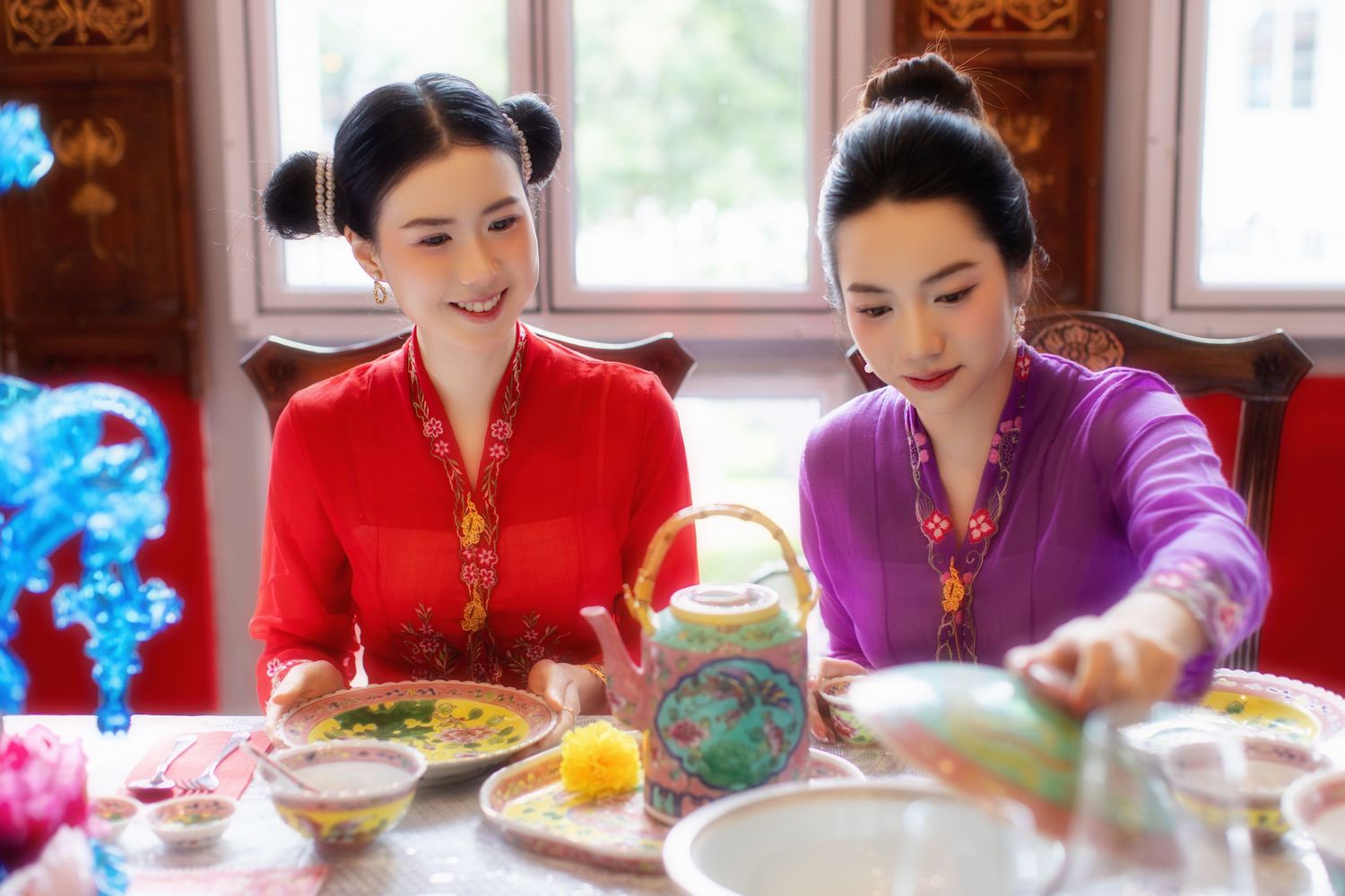 Photoshoot of two friends dressed in traditional Nyonya Kebaya  seated at dining table with traditional tea setting inside a Peranakan house