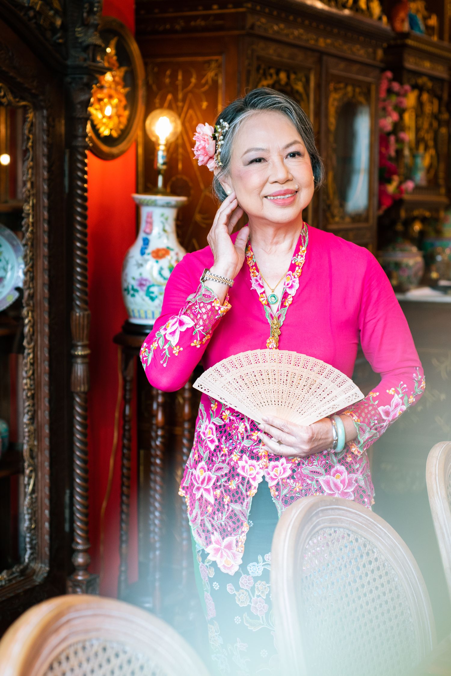 Person in traditional Asian attire wearing bright pink kebaya and holding decorative fan poses in ornate interior setting.