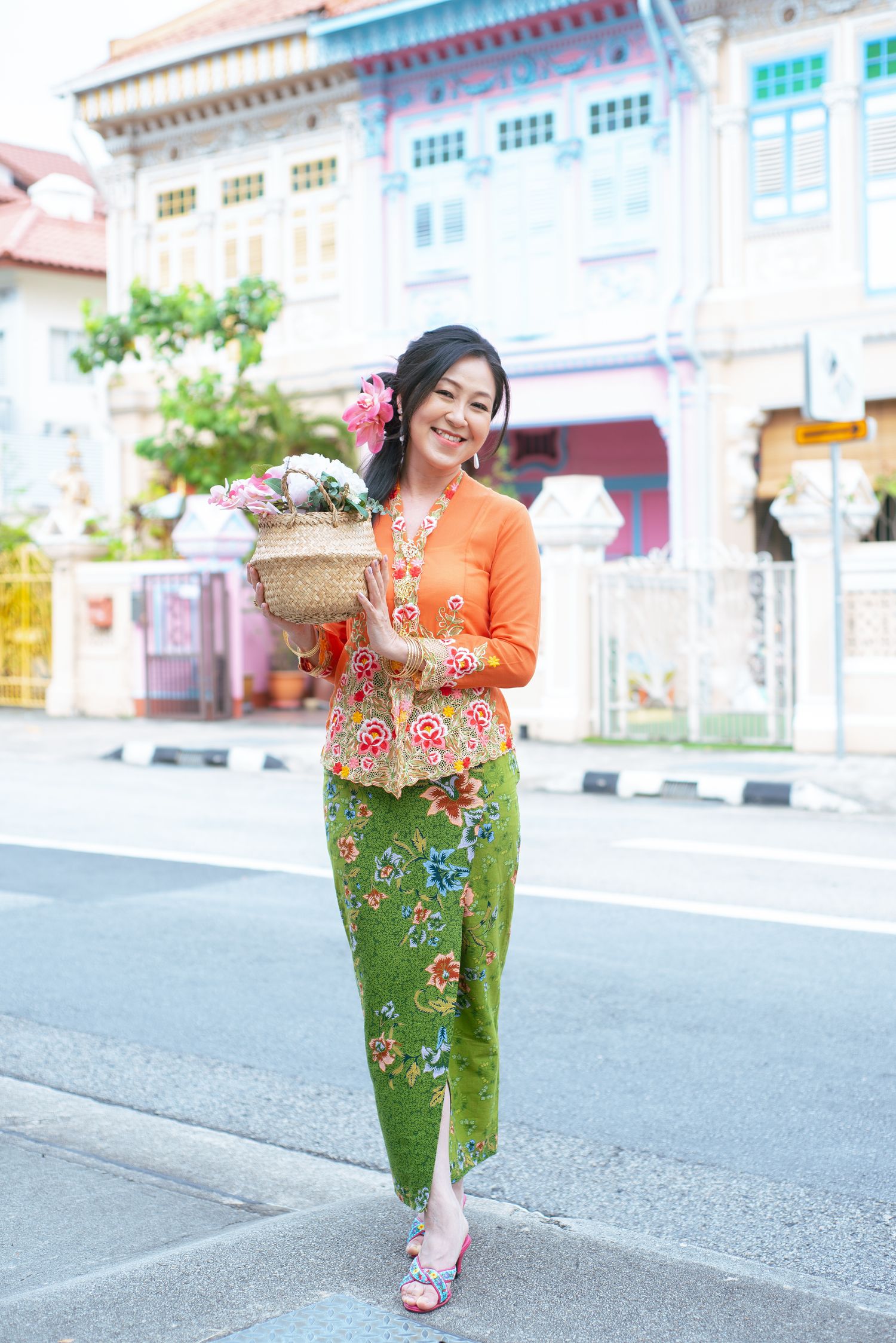 Person wearing orange kebaya top and floral sarong holding decorative basket poses on street with traditional architecture.
