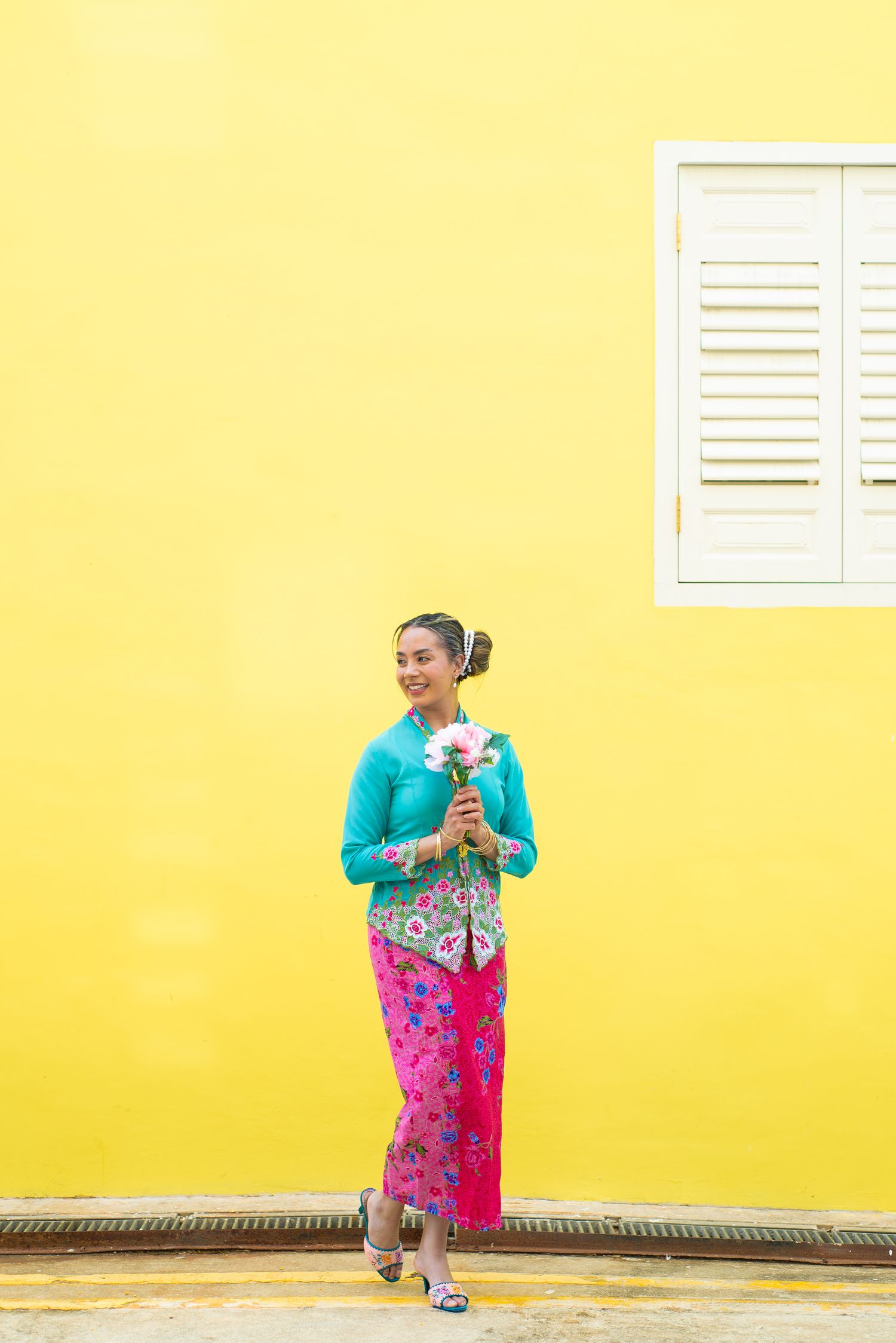 Person wearing traditional Indonesian kebaya dress with turquoise top and pink sarong stands against bright yellow wall.