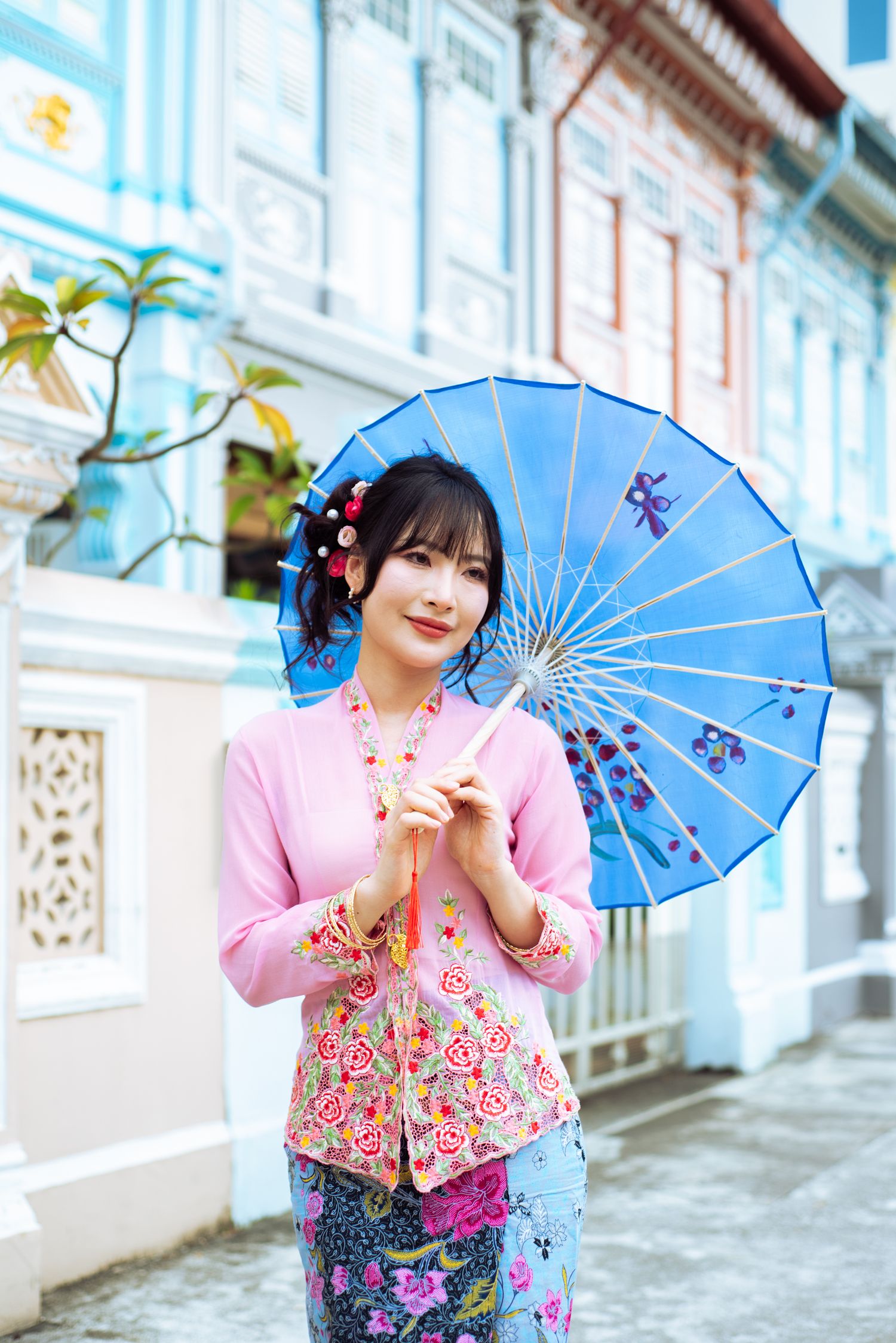 Traditional Asian fashion with a pink floral top and blue parasol against pastel colored heritage buildings.