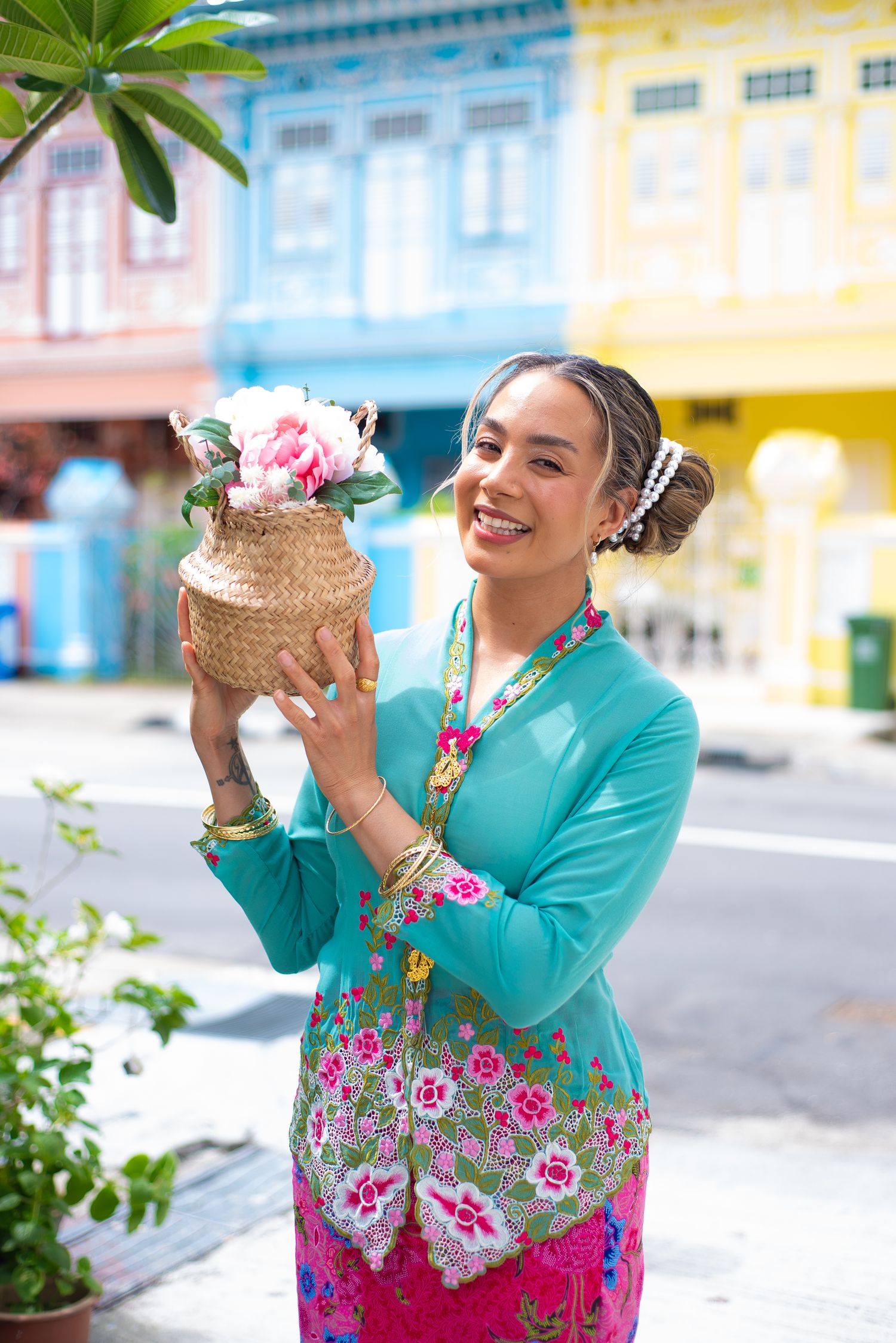 Woman in traditional Peranakan dress holding decorative vase with flowers against colorful shophouse background in Singapore.