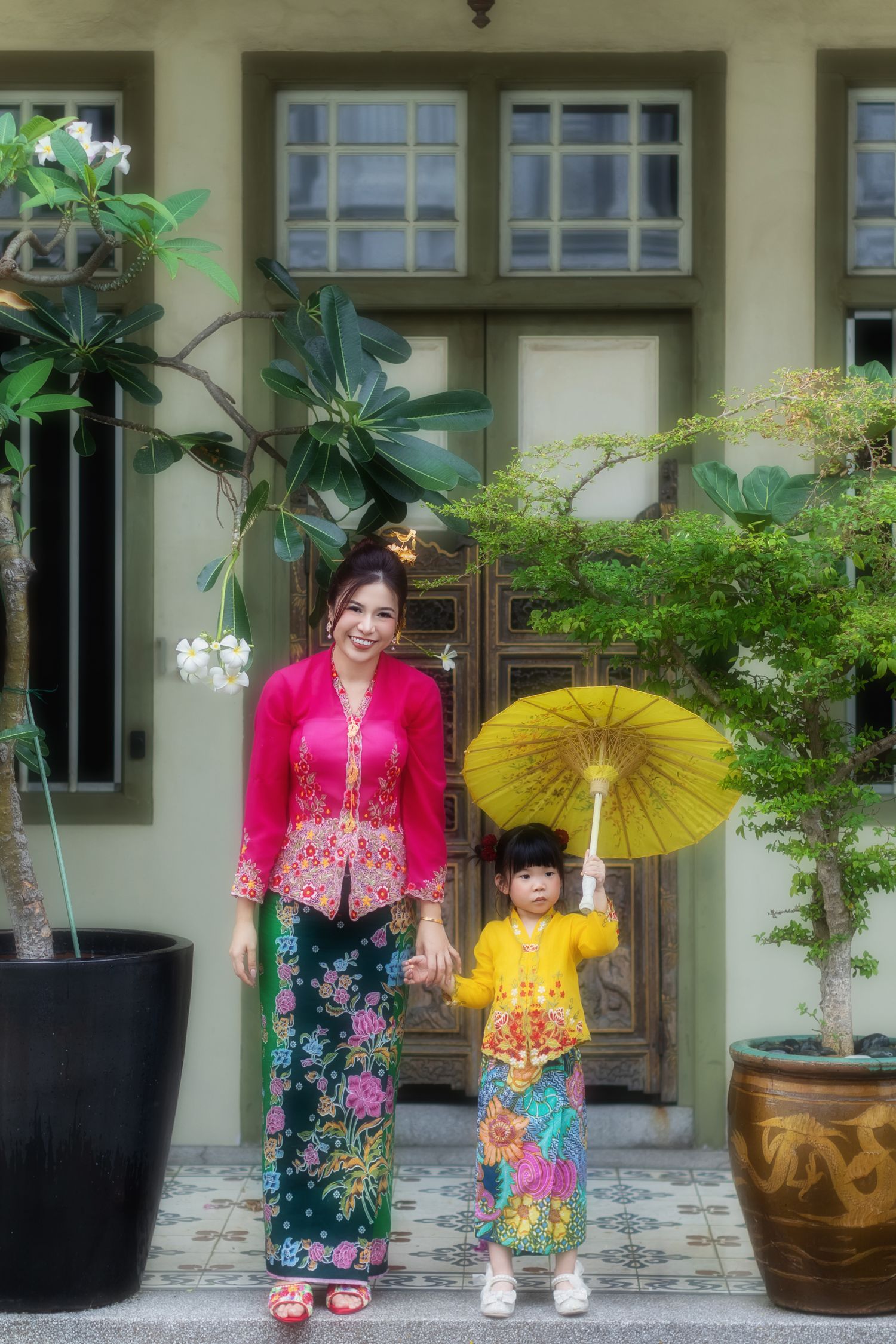 Image of a Nyonya mother and child in pink and yellow kebaya, in front of a wooden door Pintu pagar, at a Peranakan house in Joo Chiat.
