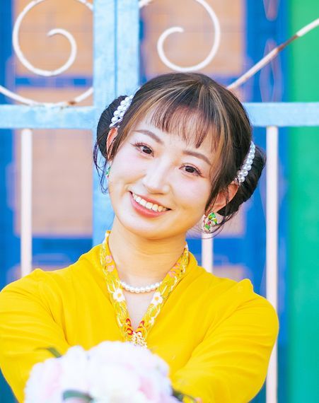 Young woman in bright yellow top smiles warmly with floral hair accessories and layered necklaces against colorful background.