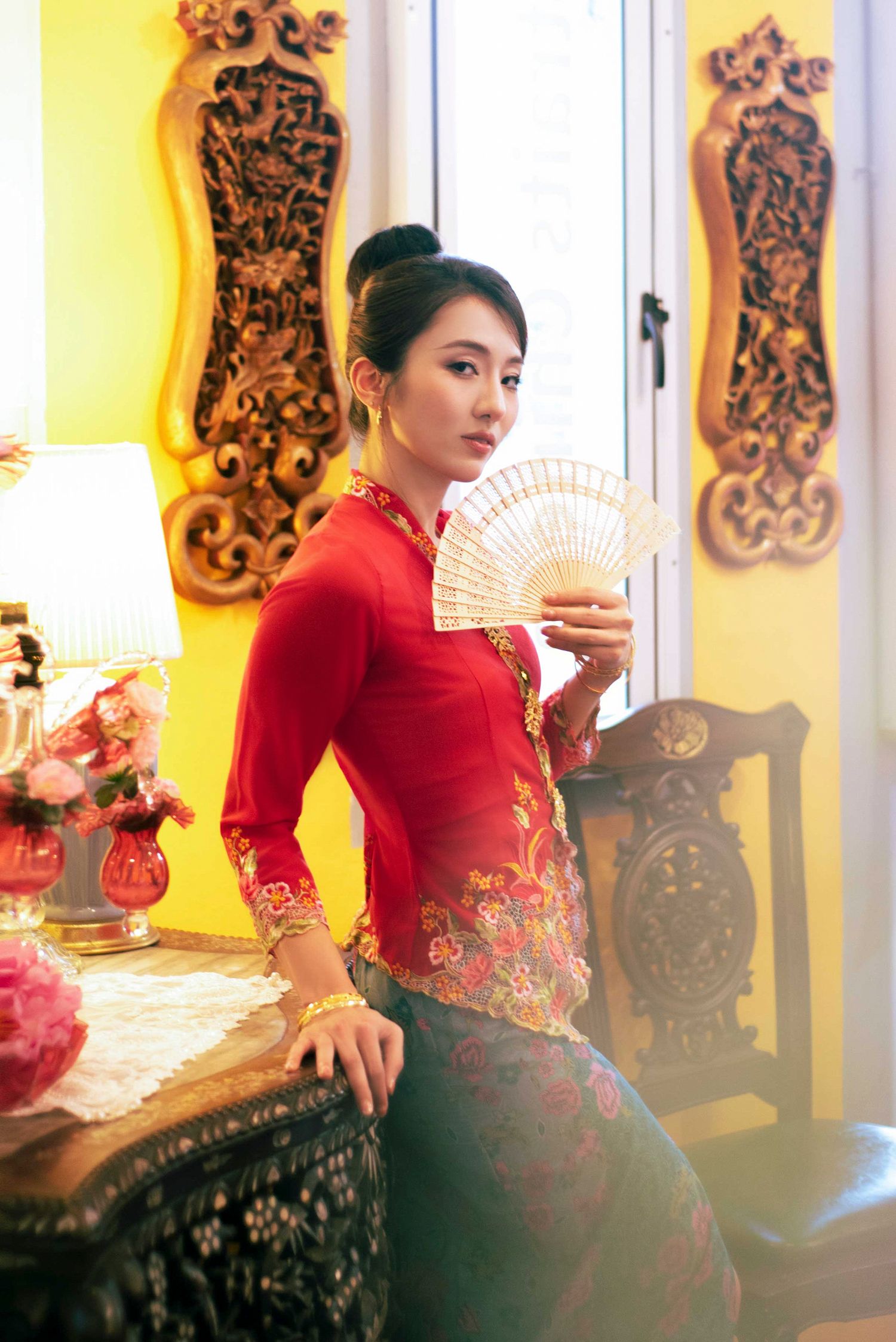 Elegant figure in traditional red Chinese dress poses with folding fan in ornate interior with carved wall panels and warm lighting.