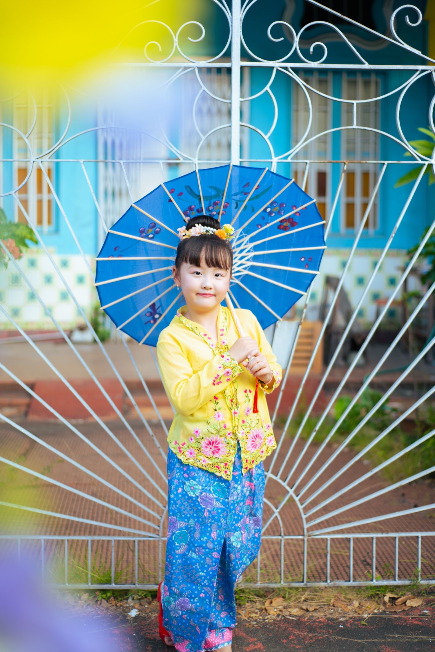 Traditional blue and yellow Asian outfit paired with a decorative blue parasol against ornate white iron gates.