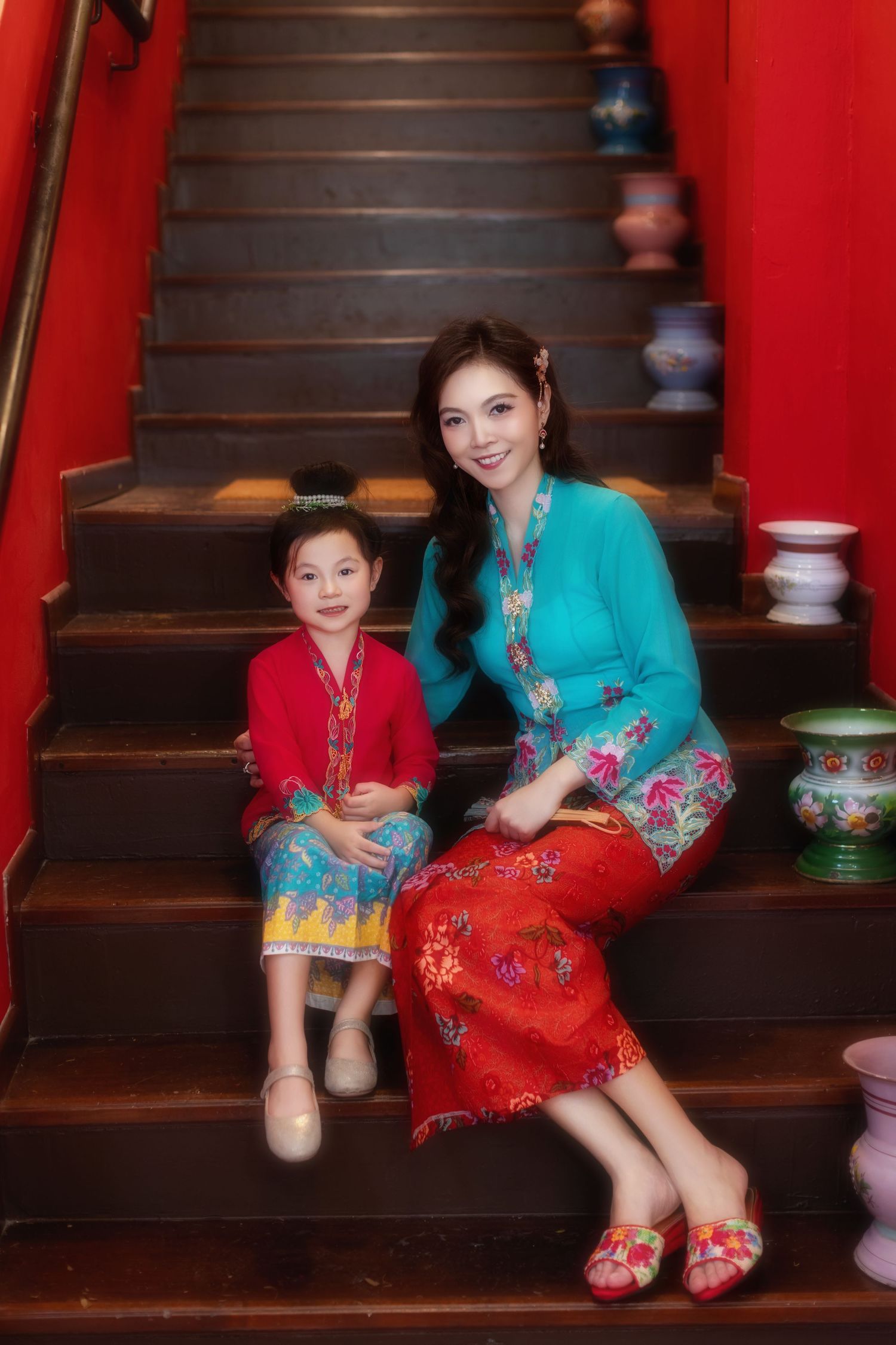 Photo of a Mother and daughter in traditional Nyonya Kebaya sitting on red-walled stairs with decorative vases.
