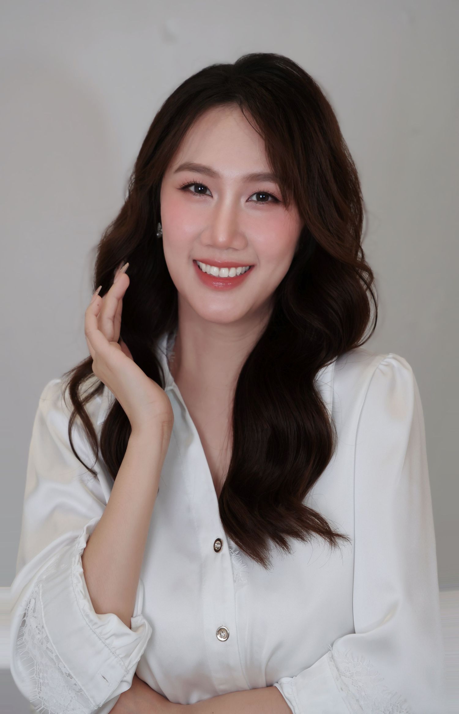 Smiling young Asian woman with long wavy brown hair wearing a white blouse, posing gracefully against a light background.