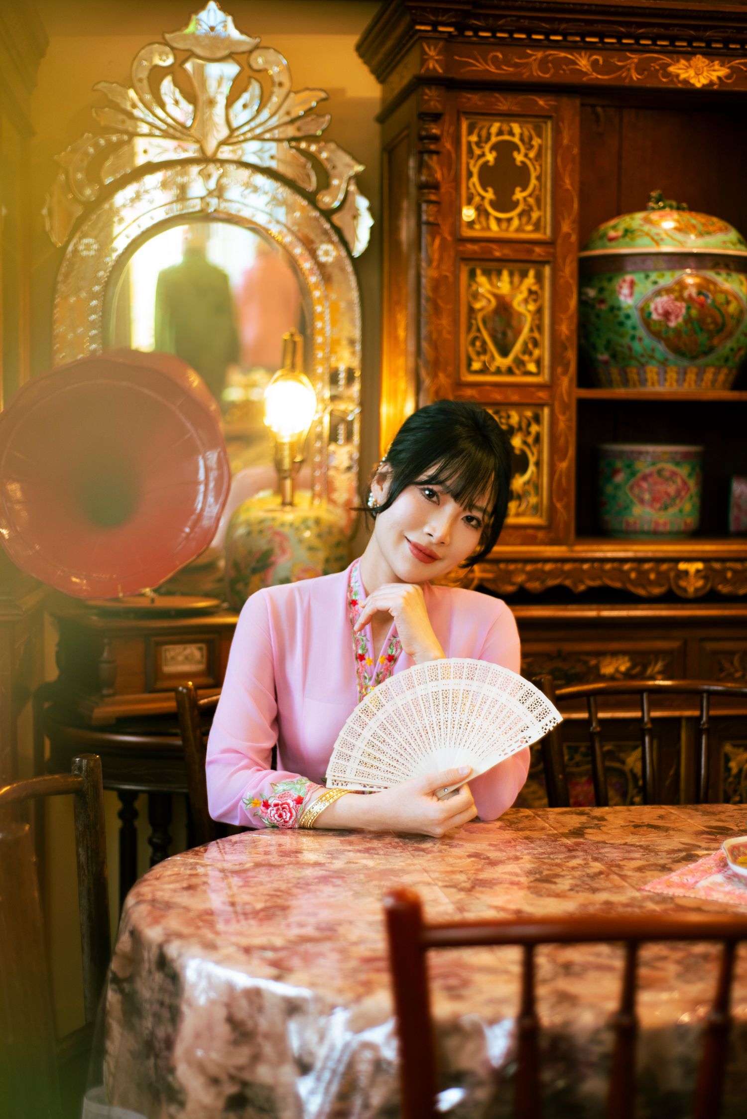 Person in a pink traditional Asian-style dress holds a decorative fan while sitting in an ornate vintage-style interior setting.