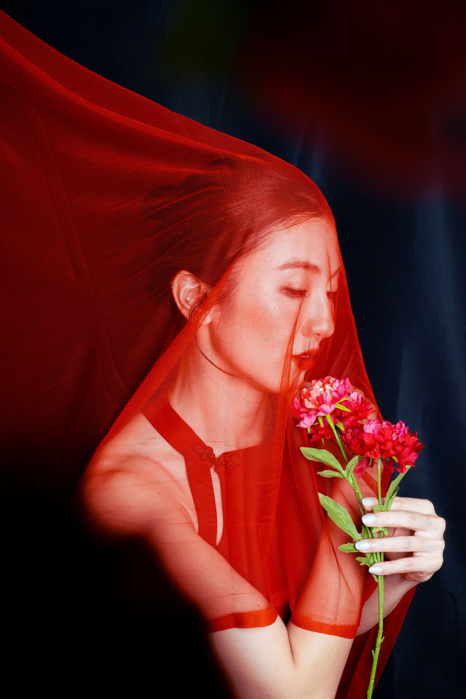 A young Asian woman in red sheer dress holds pink flowers under a flowing red veil against dark background.