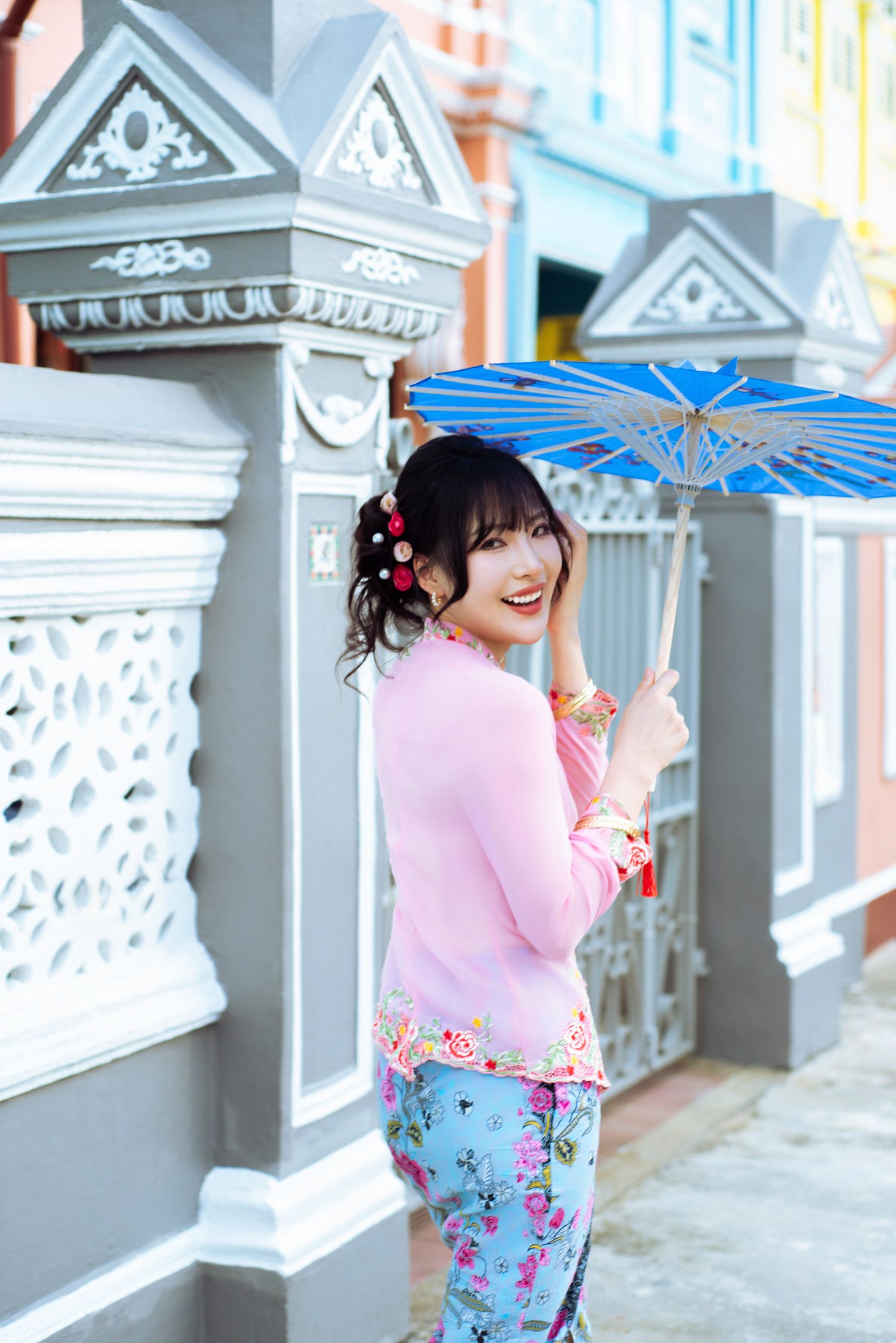 Person in pink dress with floral print poses playfully near ornate gray architectural columns with blue umbrella.