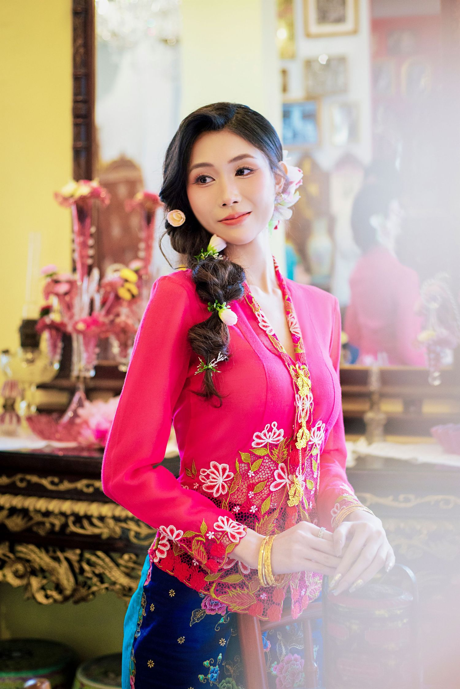 Traditional Peranakan-style dress in bright pink with golden floral embroidery photographed in an ornate interior setting.