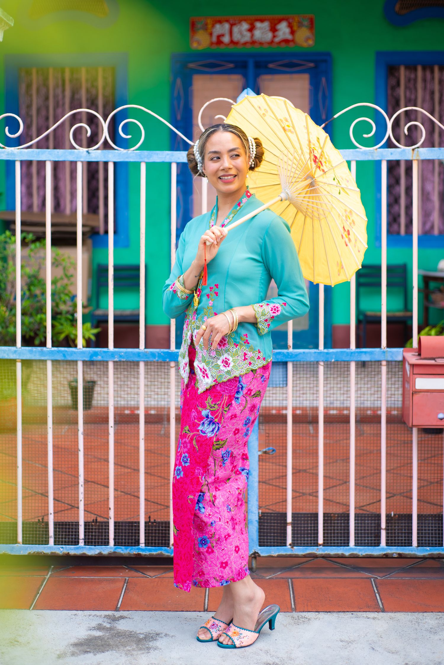 Woman in traditional Malaysian dress with turquoise top, pink sarong and yellow parasol poses by ornate metal gate.
