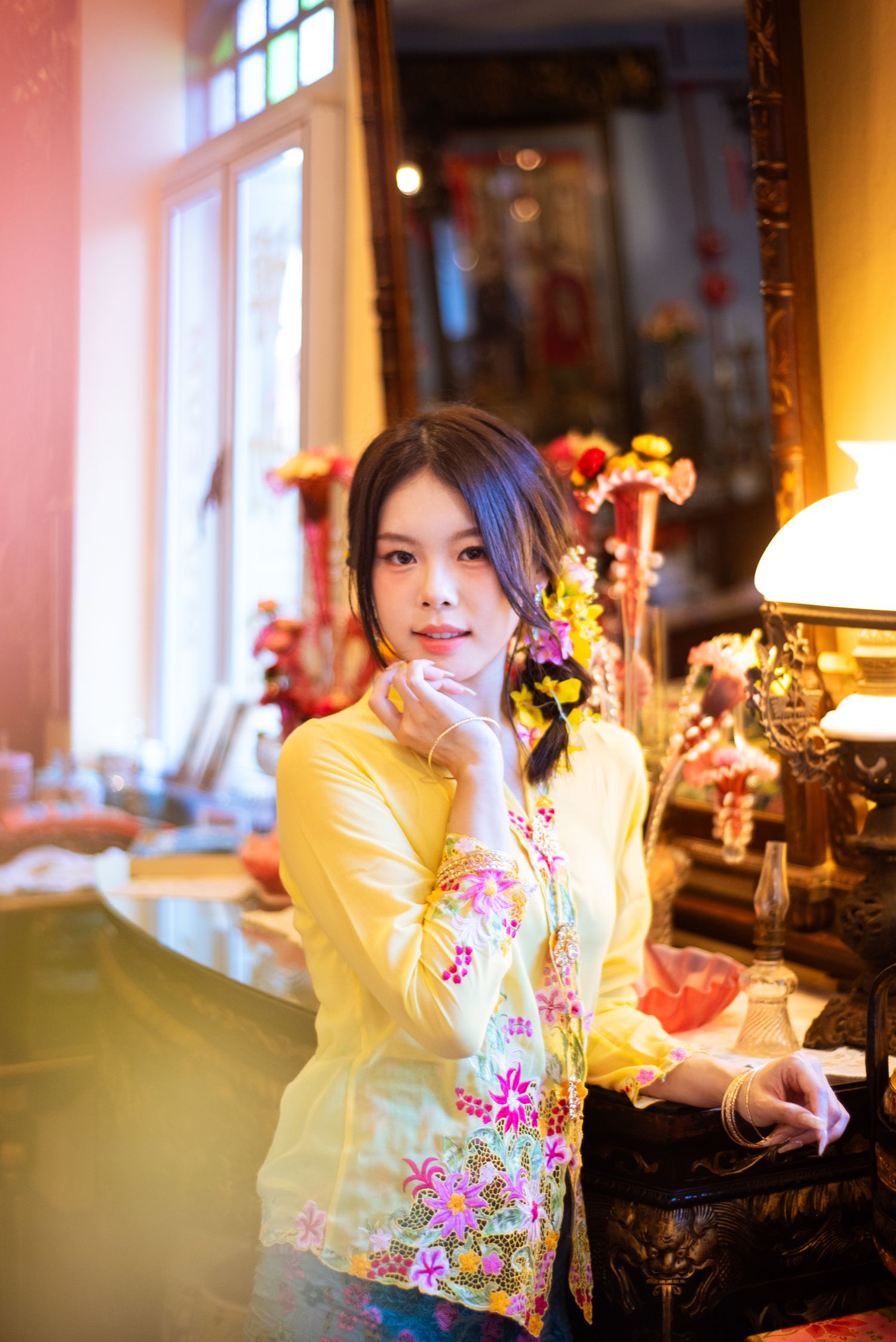 Woman in yellow and pink floral traditional Asian attire poses elegantly by a window with decorative antique furniture.