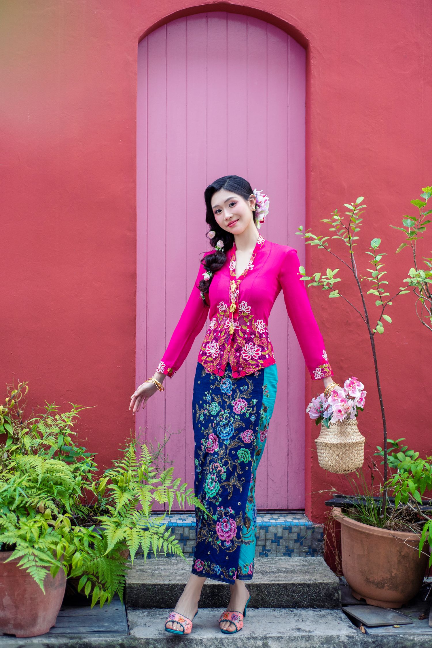 Traditional Malaysian outfit with pink kebaya and floral sarong posing in front of purple door against coral red wall.