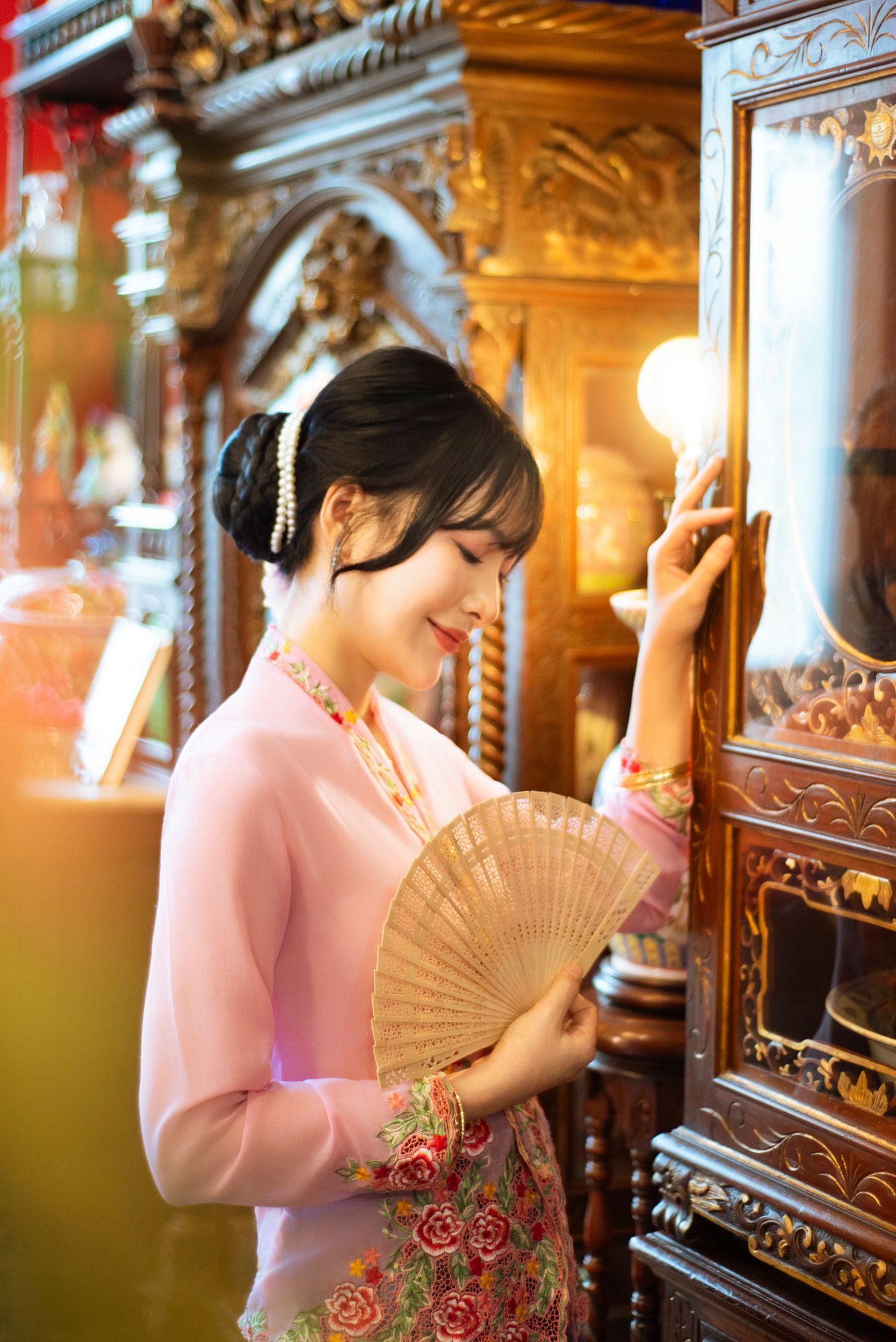 A person in a pink traditional Asian dress holds a fan while standing near ornate wood carvings in a warm-lit interior.