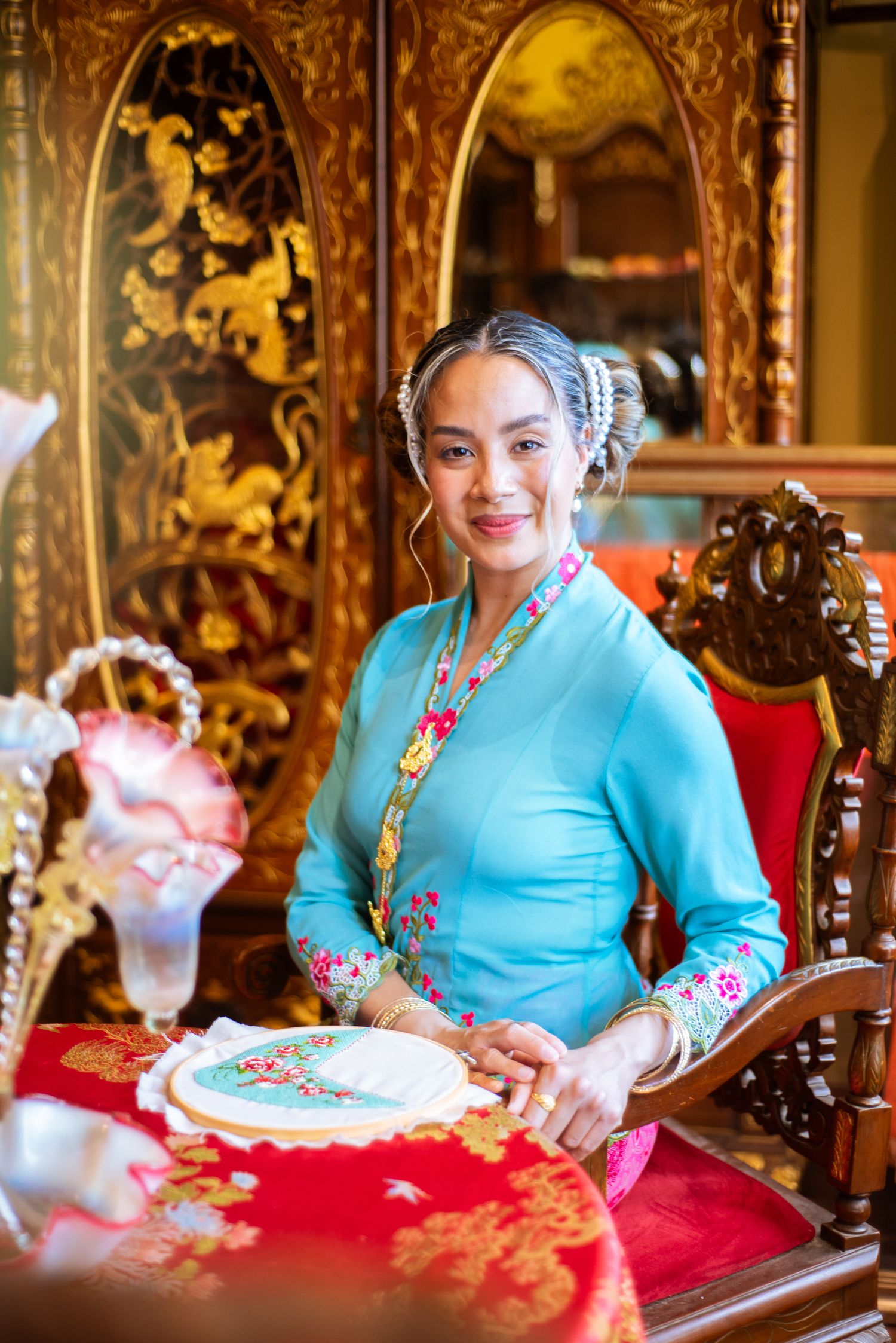A person in a turquoise embroidered dress sits at an ornate wooden table with red fabric in a vintage-styled setting.