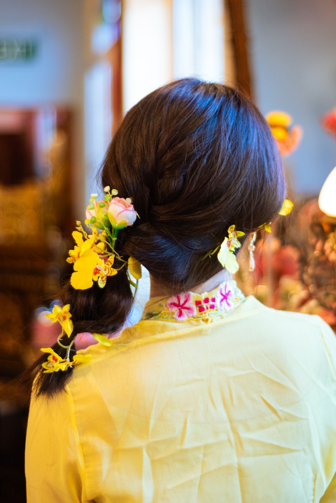 A dark-haired person wearing a yellow top adorned with colorful flowers woven into their braided hair.