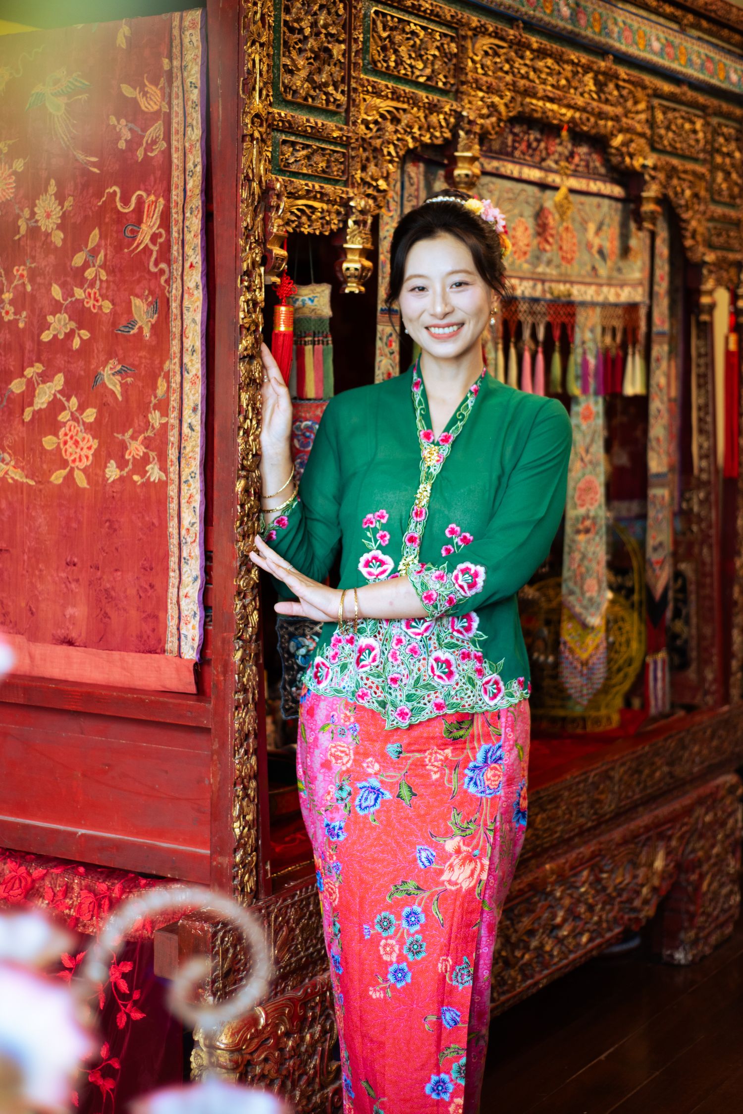 Woman in traditional green and pink floral Peranakan kebaya outfit smiling inside ornate red and gold heritage building.