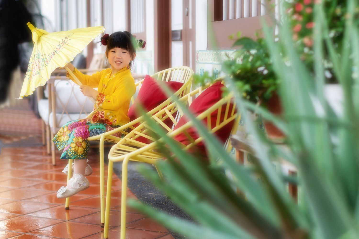 Photo of a girl in a yellow Nyonya Kebaya sits on a porch with colorful chairs and tropical plants visible, holding a yellow umbrella.