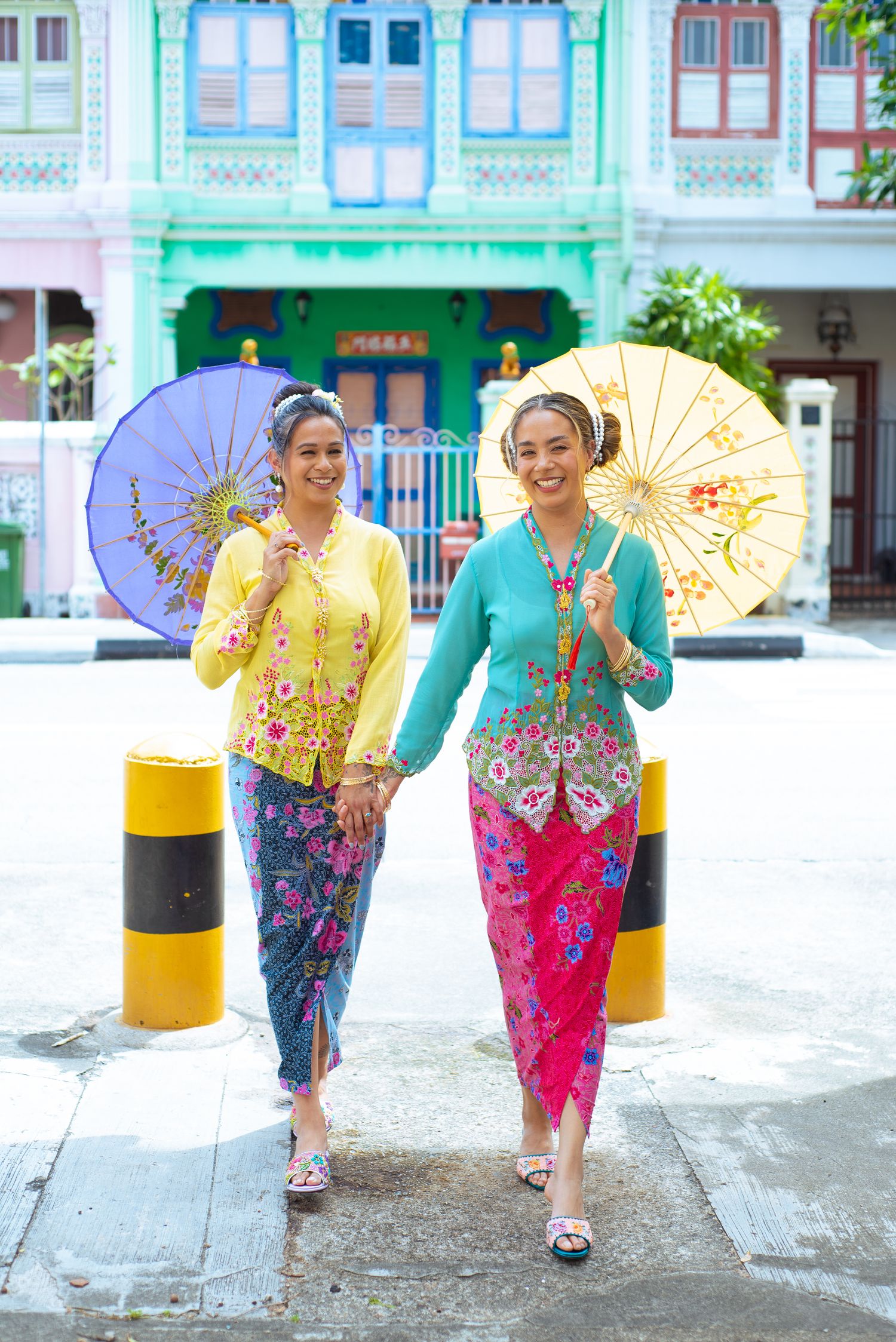 Two women in traditional Peranakan clothing with colorful parasols walk along a heritage shophouse street in Singapore.