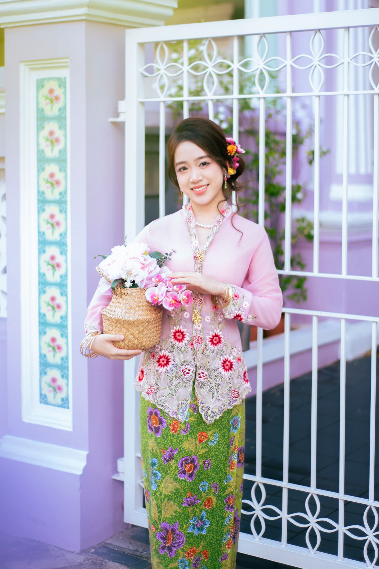 Woman in traditional Peranakan Nyonya kebaya dress holding flower basket stands by ornate white gate and purple wall.