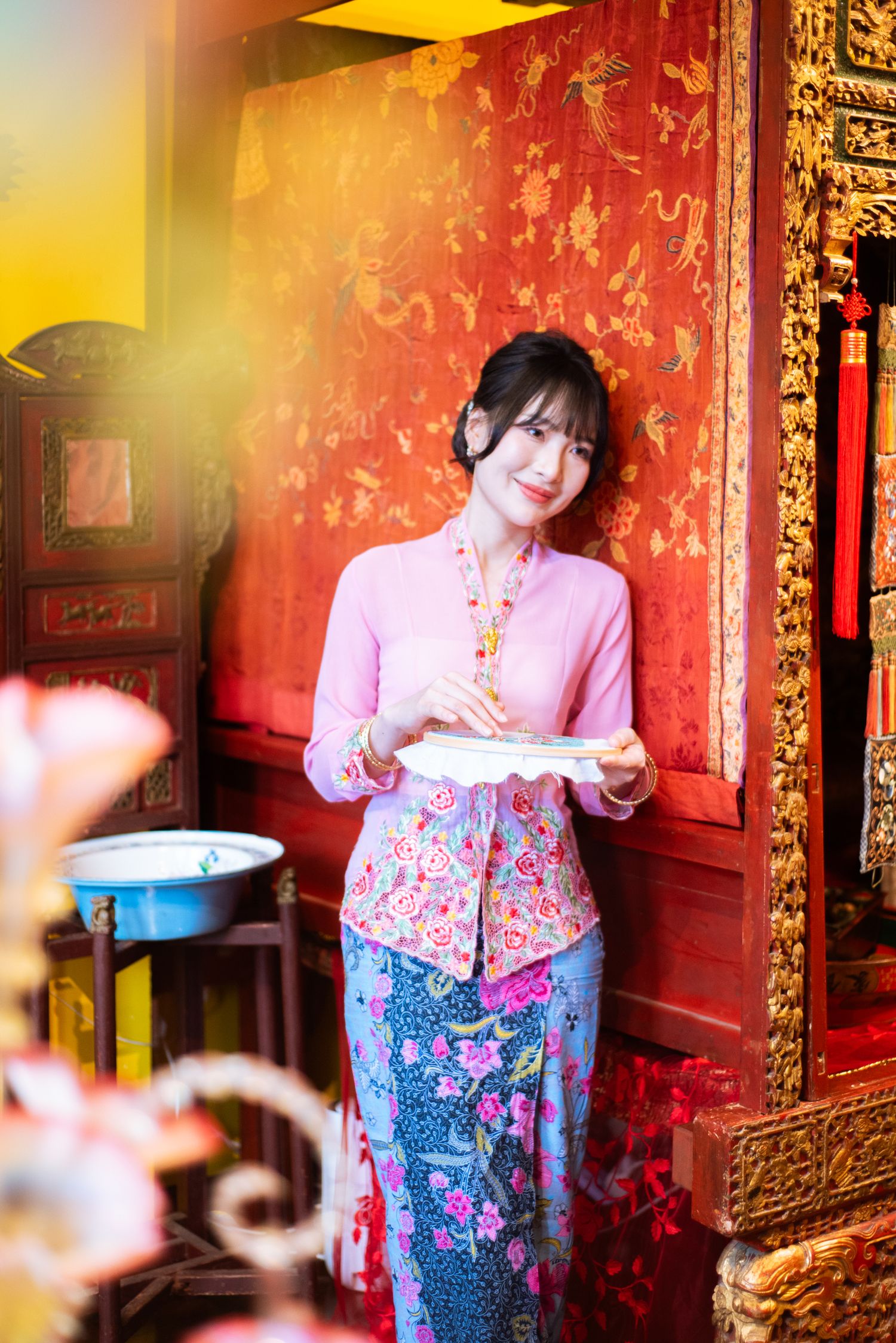 Traditional Chinese decor with red walls and gold trim serves as backdrop for someone in pink blouse and floral blue dress.