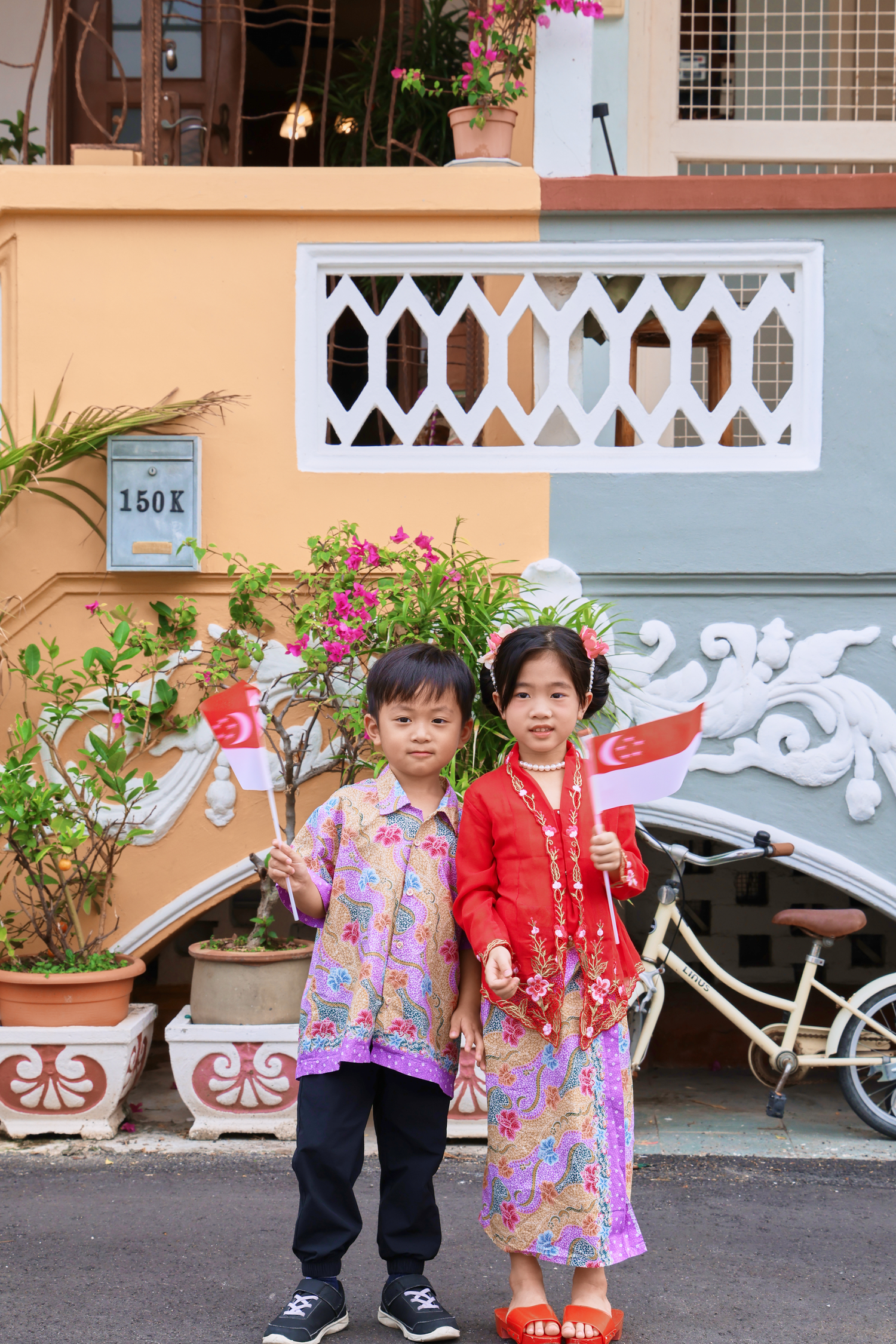 Two children in traditional Malay attire holding Singapore flags pose in front of a colorful heritage shophouse.