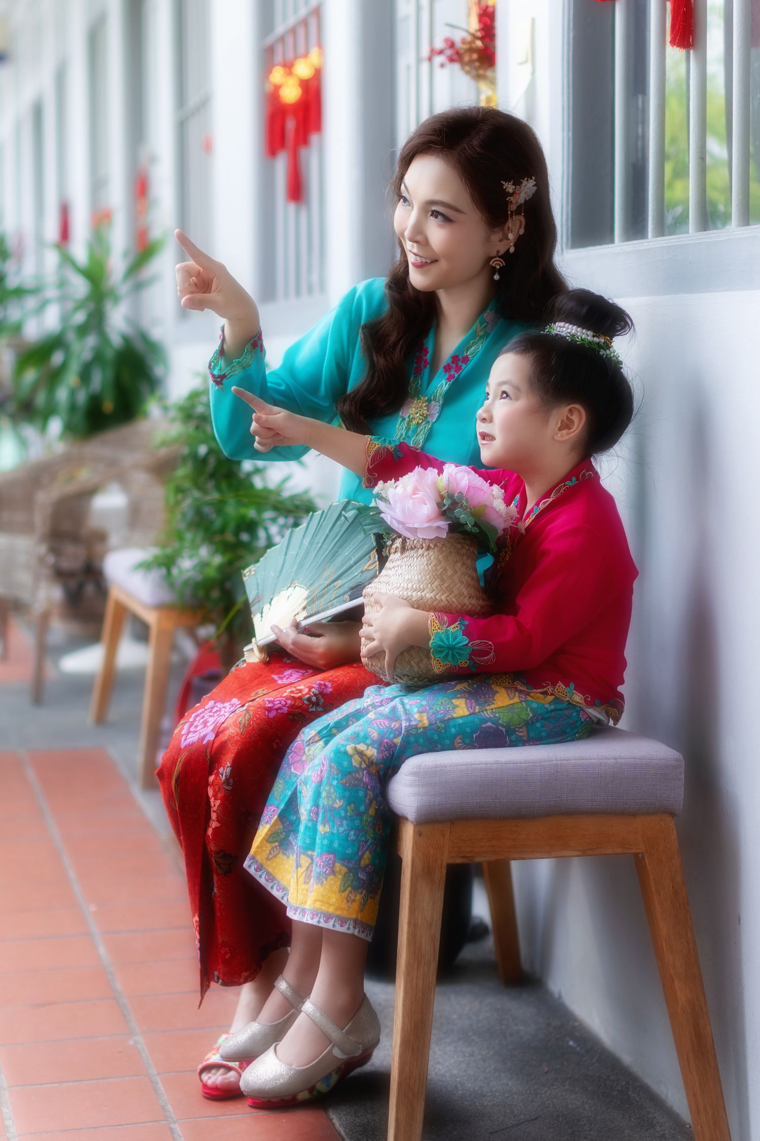 Photoshoot of a mother and daughter in traditional Nyonya Kebaya sitting on chairs by a wall carrying a basket of flowers.