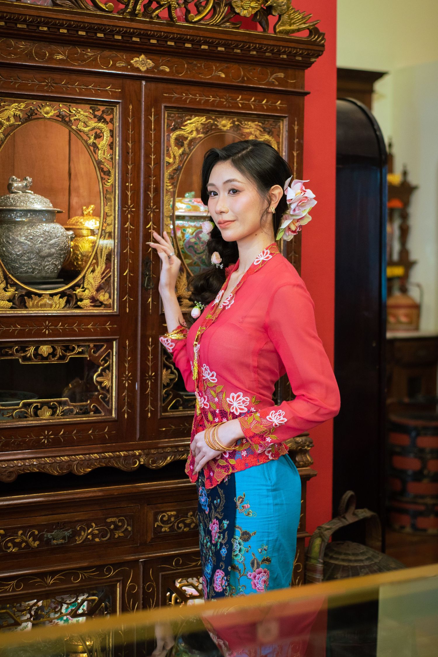 Traditional kebaya dress in red and blue photographed against an antique wooden cabinet with gold details.
