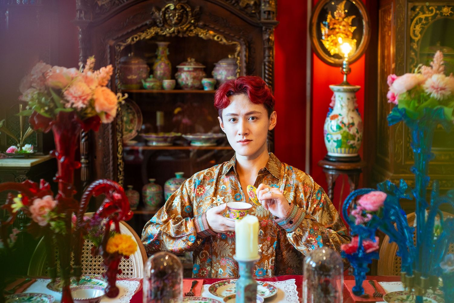 Young man with red hair in ornate room, wearing silk shirt, sitting at decorated table with candle and flowers.