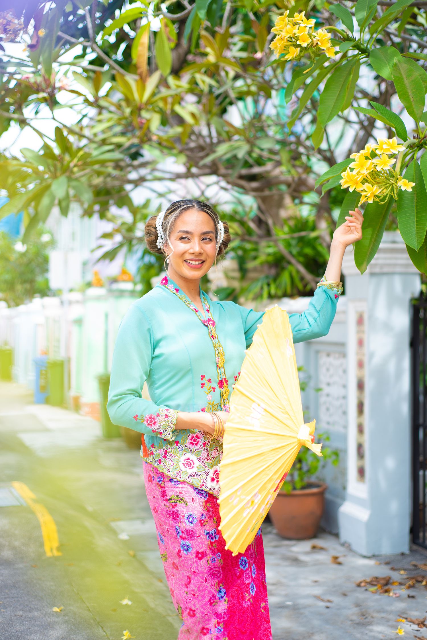 A person in traditional Southeast Asian attire holds a yellow paper parasol while standing next to tropical plants.
