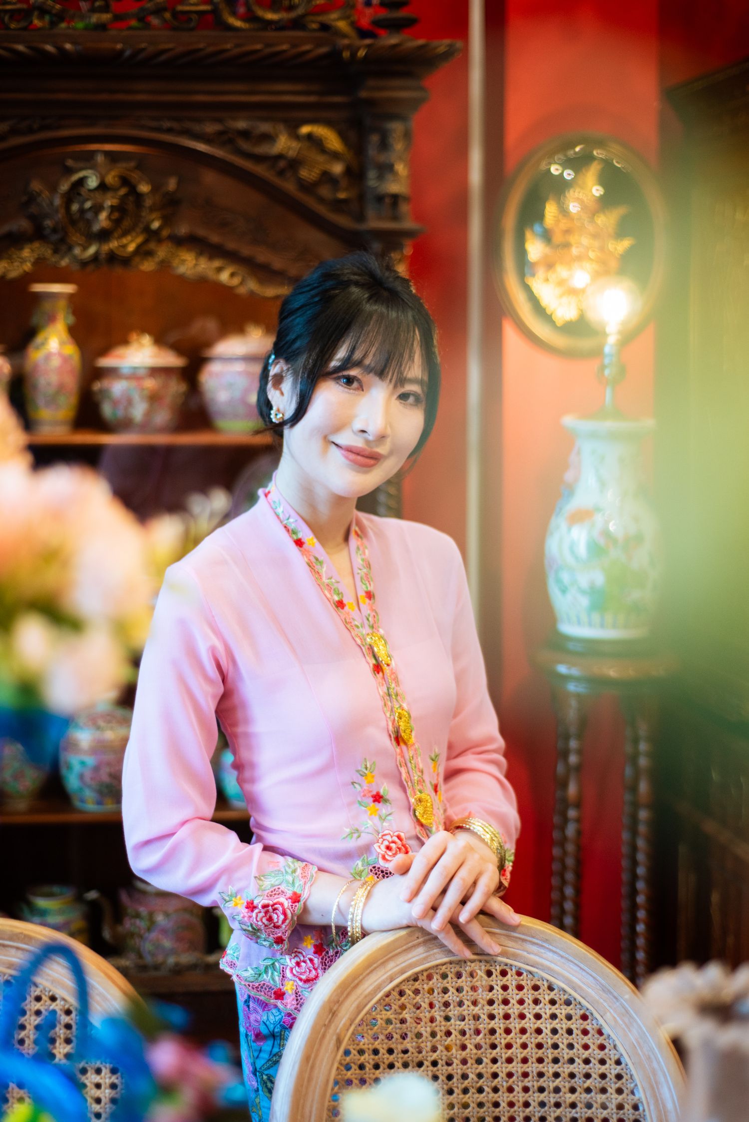 A person in a pink sweater and floral accents poses in a vintage-style interior with ornate furniture and decorative vases.