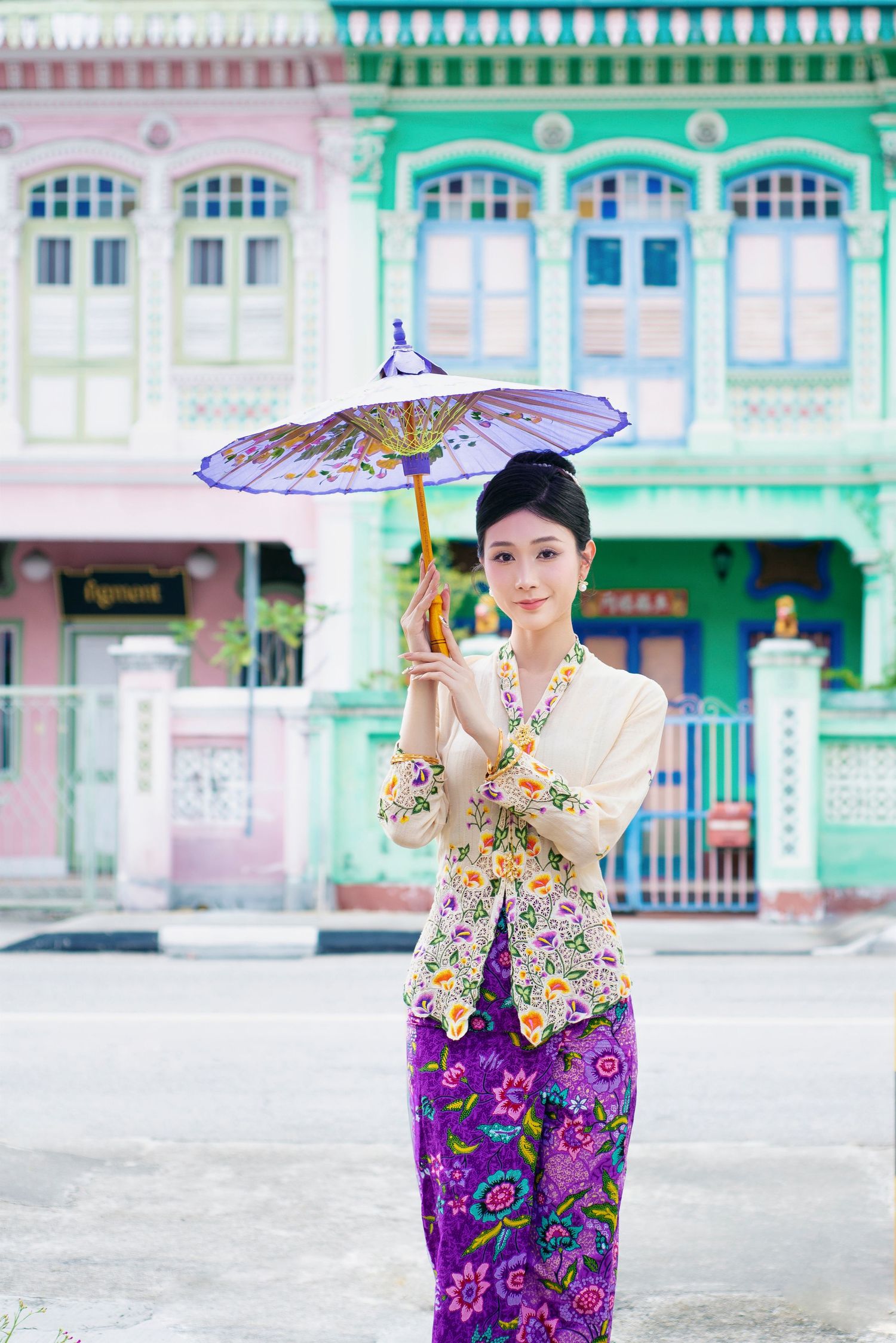 Traditional Southeast Asian attire with floral patterns and decorative umbrella against colorful heritage shophouse facade.