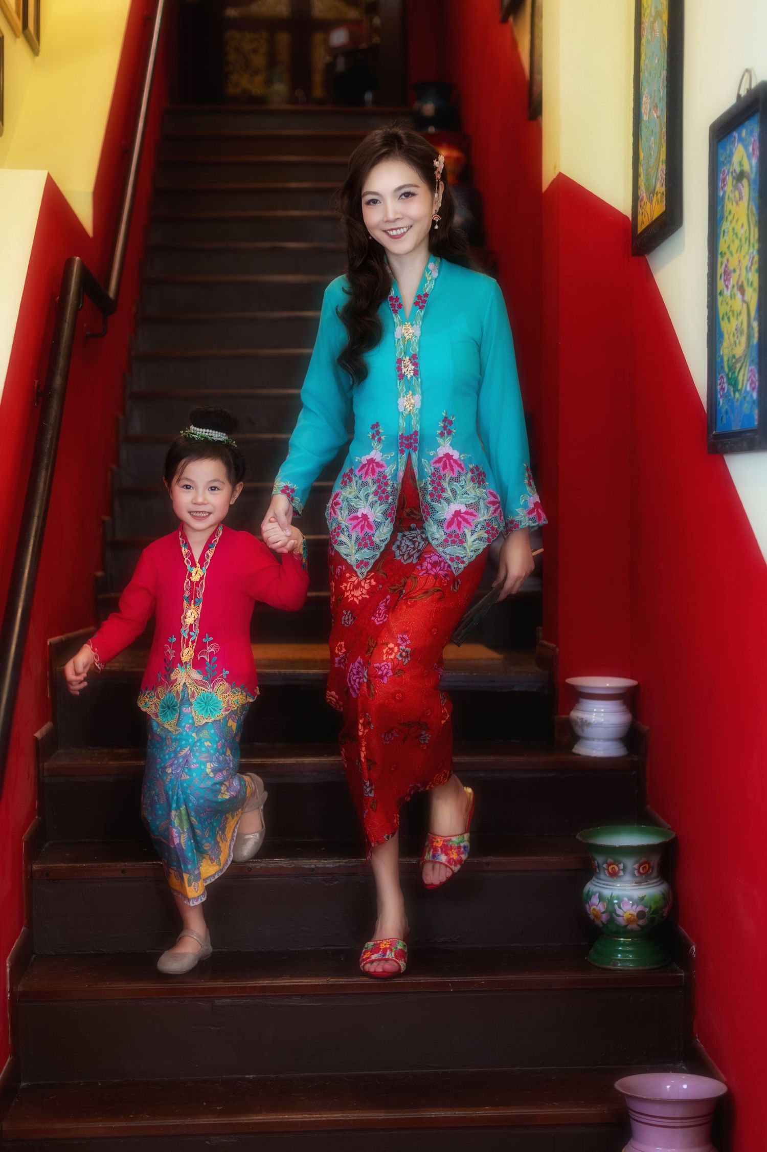 Photo of a Mother and daughter in traditional Nyonya Kebaya descending the red-walled stairs with decorative vases.