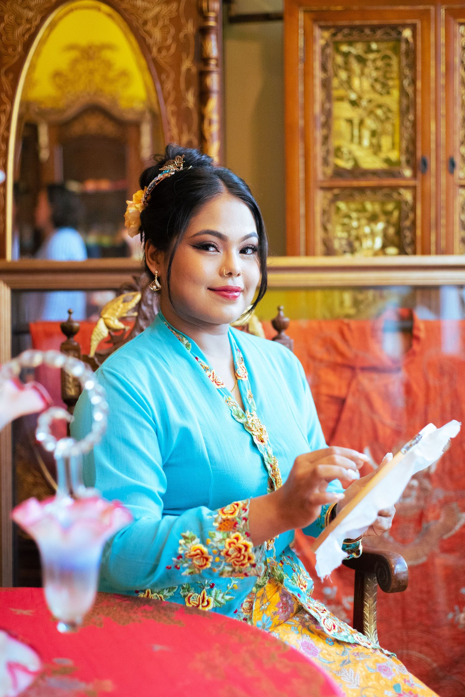 A person in traditional Myanmar dress with turquoise jacket and gold accessories sits at a red table holding papers.