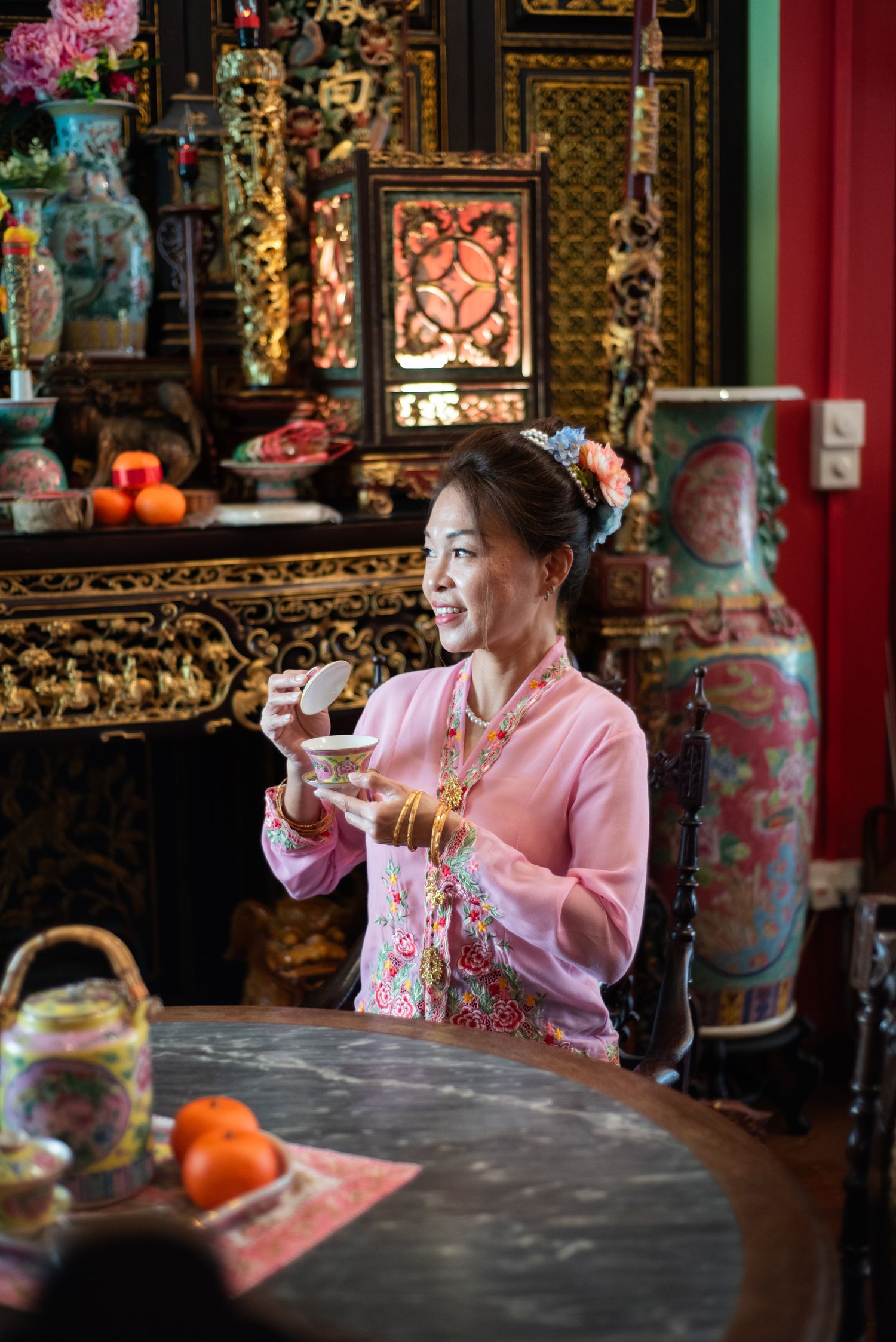 Someone enjoys tea in traditional pink attire at an ornate Asian-style interior with decorative vases and lanterns.
