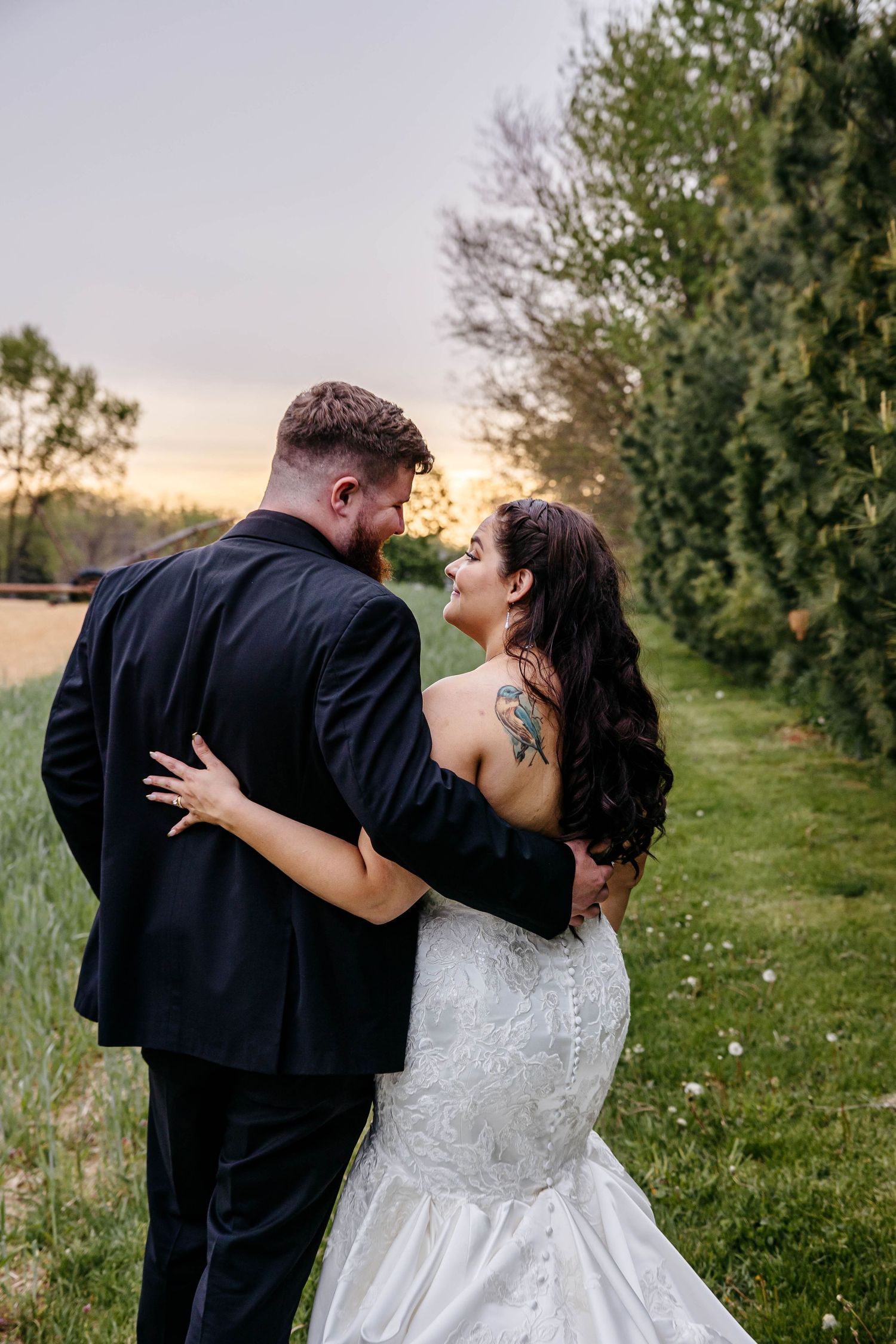 A newly married couple embraces in a romantic outdoor setting at sunset with trees and greenery in the background.