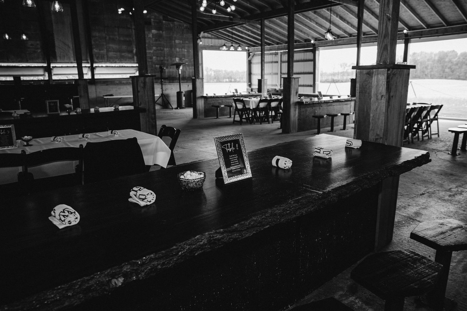 Black and white interior view of an empty rustic bar counter with seating and windows at a lakeside venue.