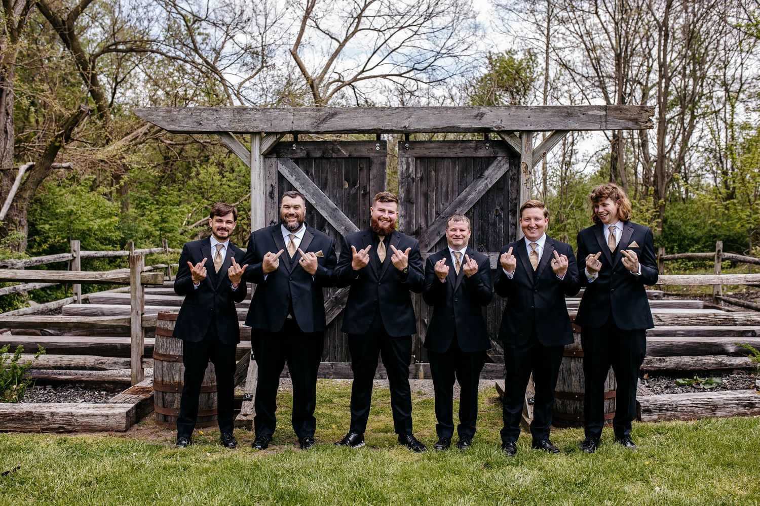 Groomsmen in black suits stand together posing in front of a rustic wooden structure and amphitheater seating.