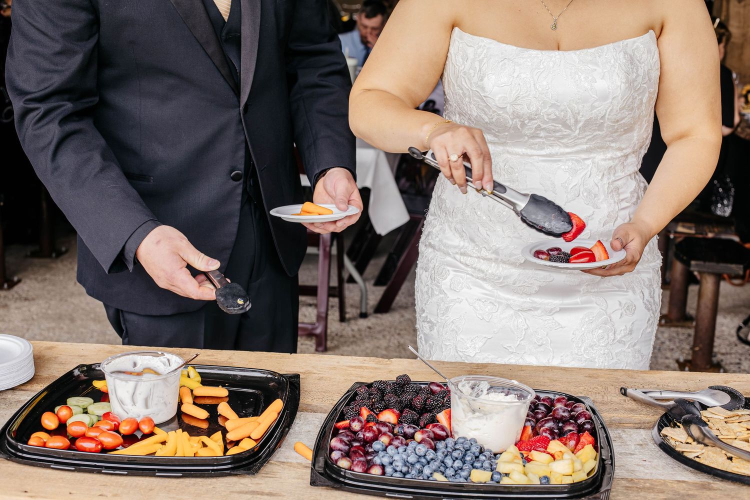 People arrange colorful fresh fruits and appetizers on black serving platters at an elegant catering event.