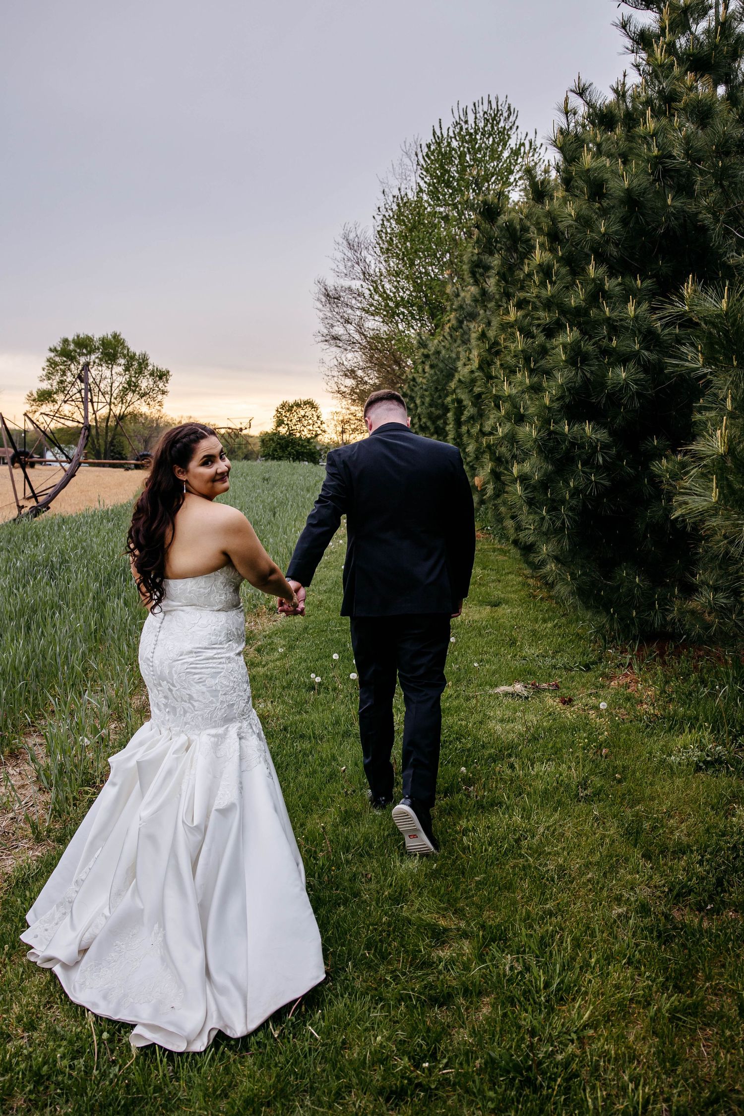 A couple in wedding attire walk hand in hand along a grassy path at sunset with evergreen trees beside them.