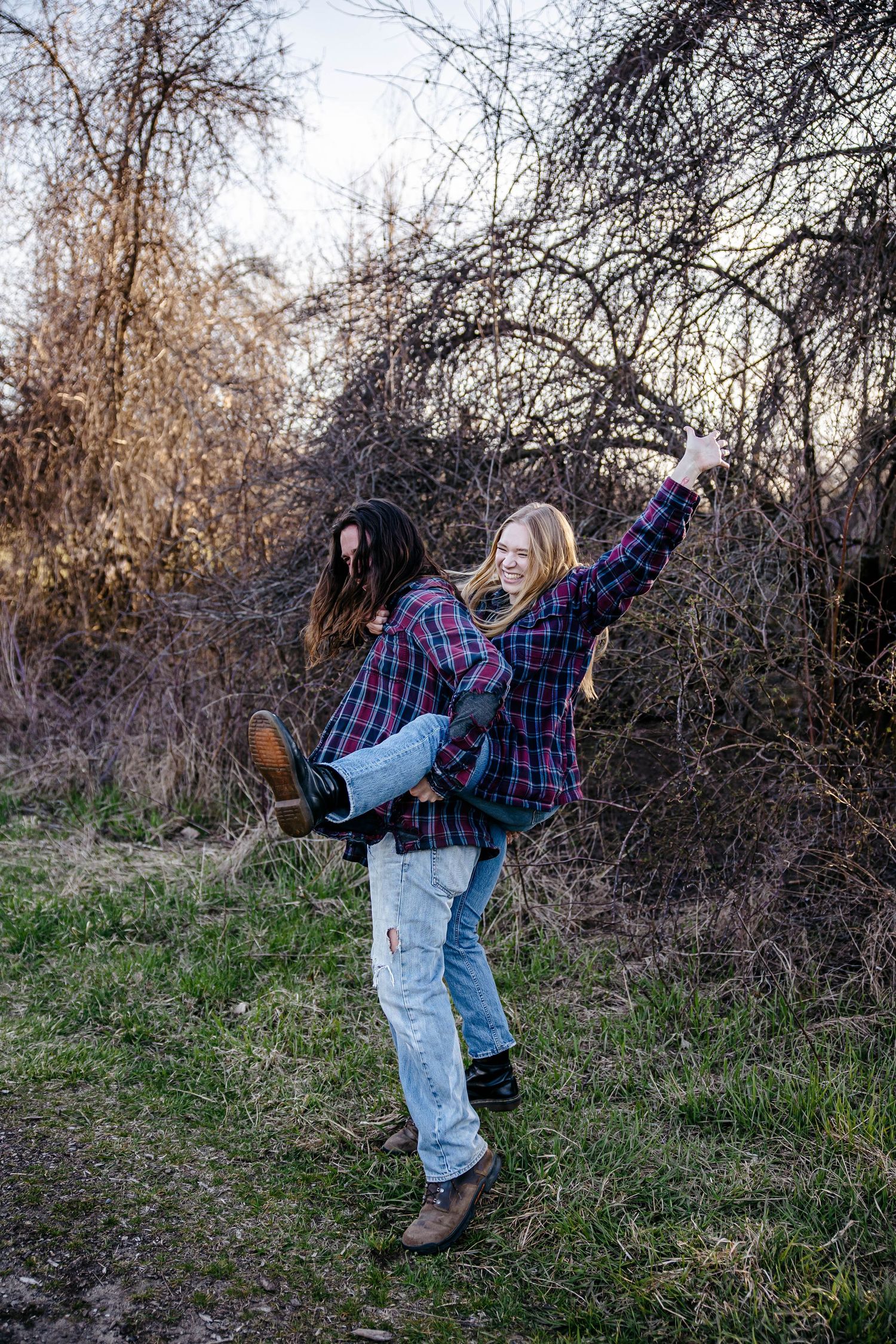 Two people playfully kick their legs up while posing together in a wooded area on a winter day.