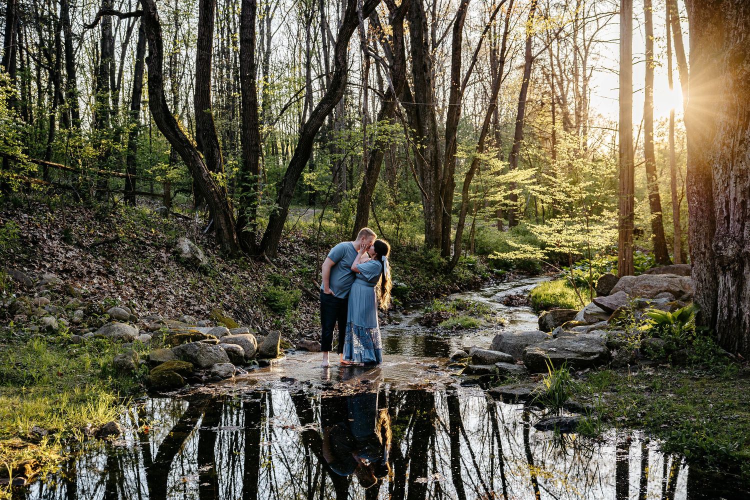 Two people stand embracing on rocks beside a tranquil forest stream with spring foliage and sunlight filtering through trees.
