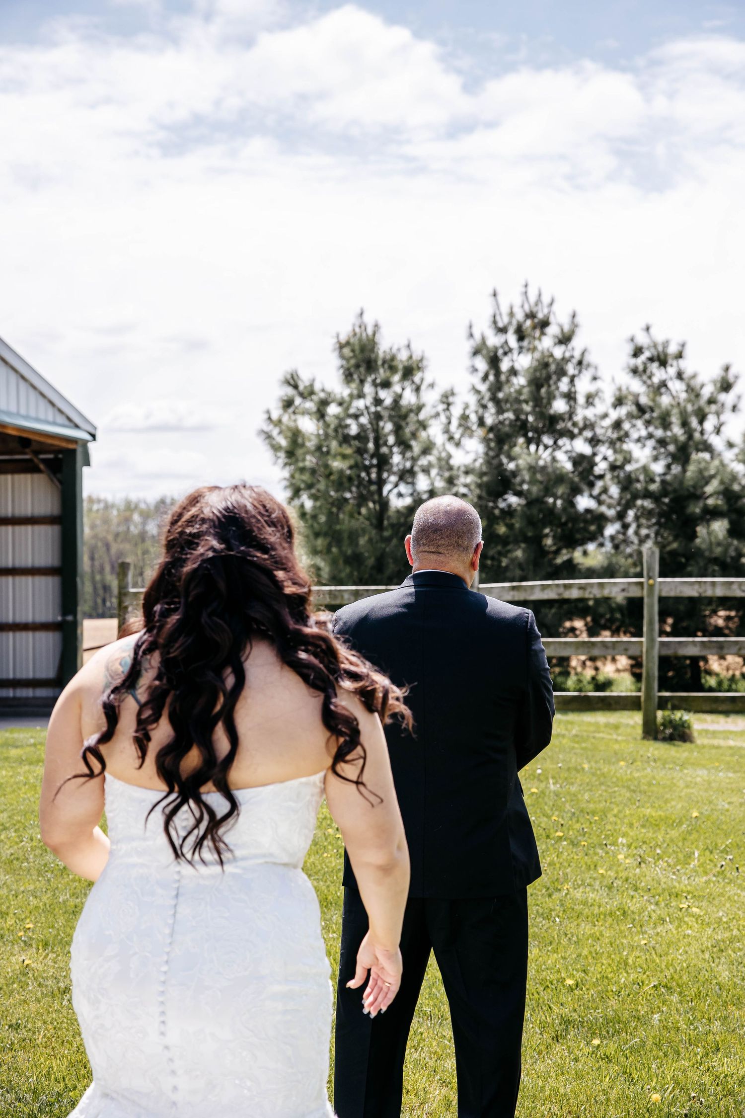 A bride in a white dress approaches her partner from behind during a first look moment in a sunny outdoor setting.