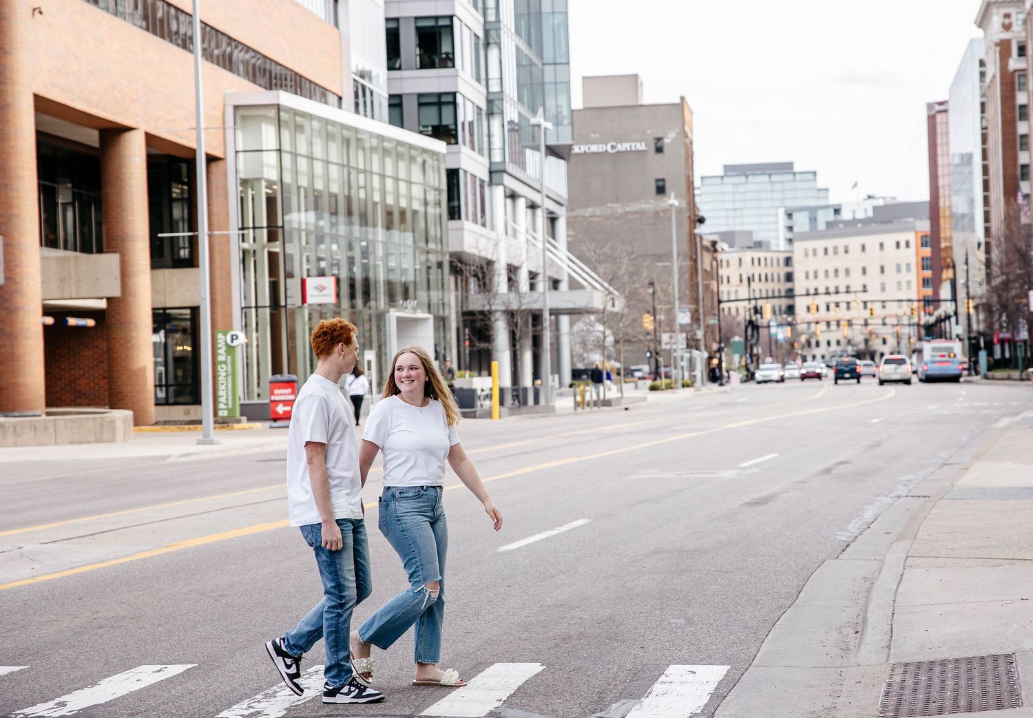 Two people cross a quiet urban street with modern buildings and storefronts in the background.